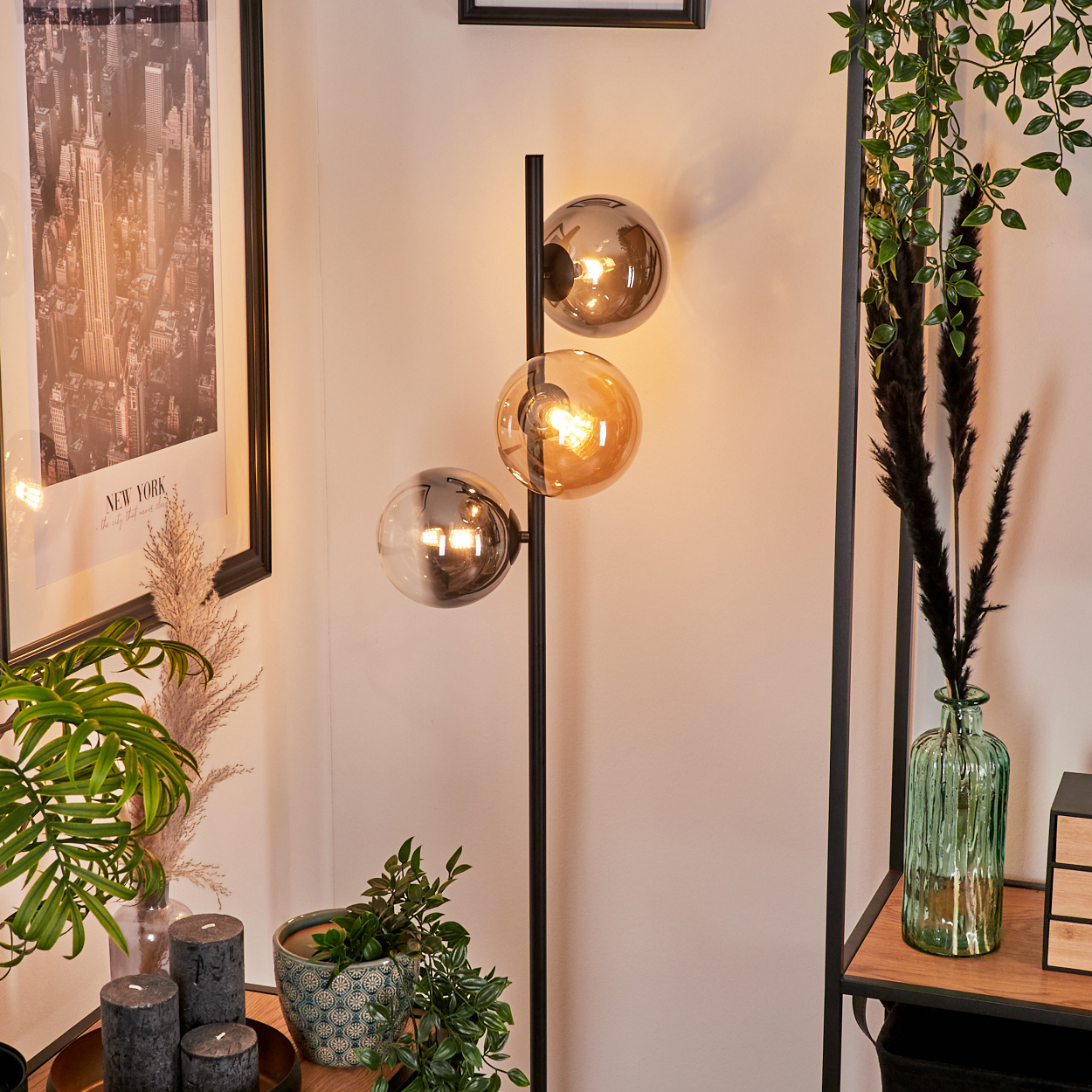 Schwarze Stehlampe mit drei Glaskugelschirmen, Pflanzen und einem Poster an einer weißen Wand.