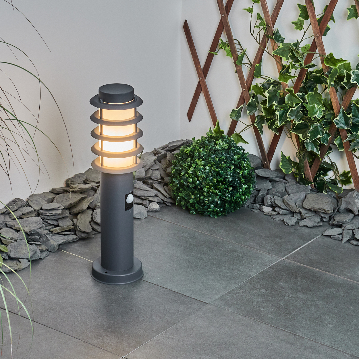 Graue Außenlampe mit Spiraldesign. Sie steht auf einer Steinterrasse mit einem grünen Busch und Spalier.