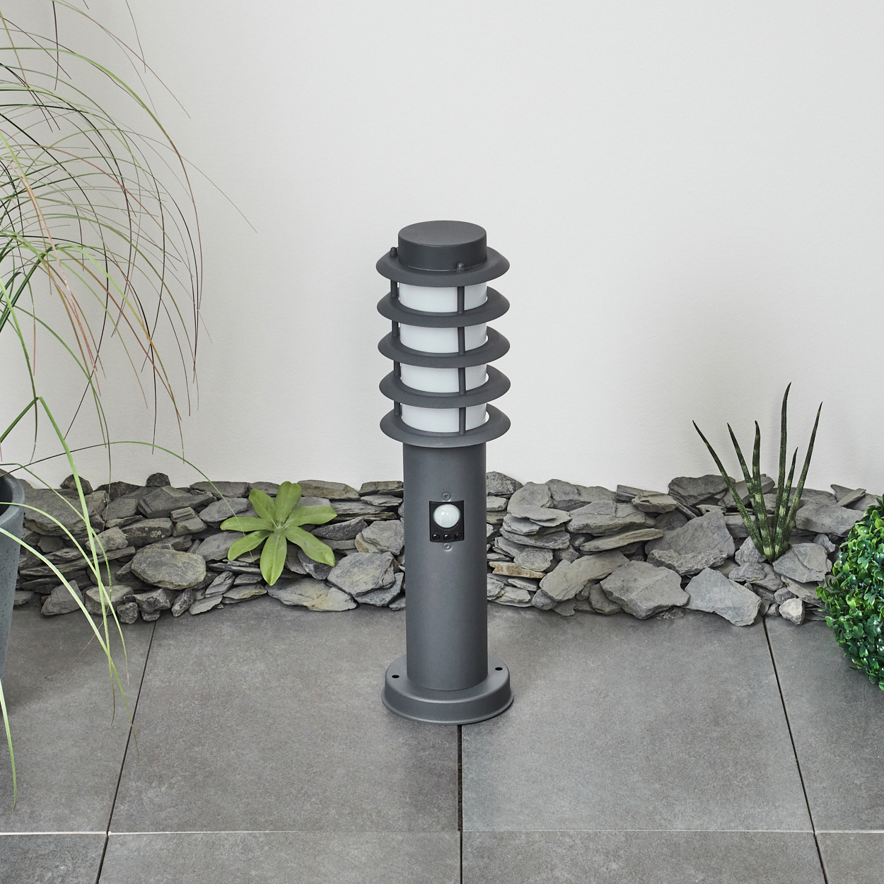 Eine Außenlaterne vor dem Hintergrund von Steinen und Pflanzen. Die Lampe ist dunkelgrau mit weißen Lichtern.