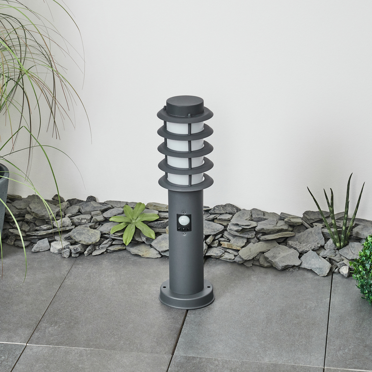 Eine Außenlaterne mit Sensor, vor einem Hintergrund aus Felsen und Grün. Die Lampe ist dunkelgrau.