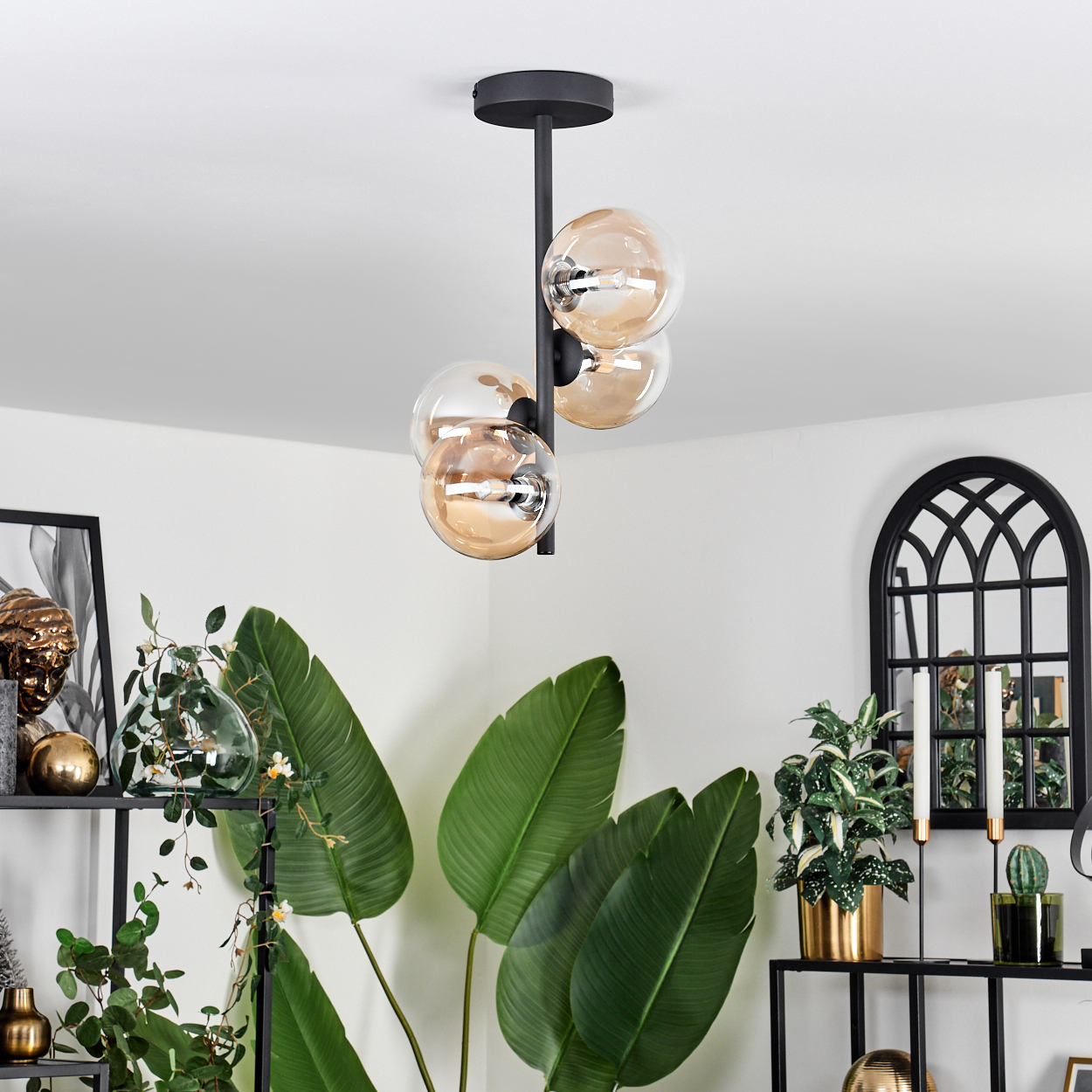 Deckenlampe mit vier bernsteinfarbenen Glaskugeln. Schwarzer Rahmen. Grüne Pflanzen im Hintergrund.
