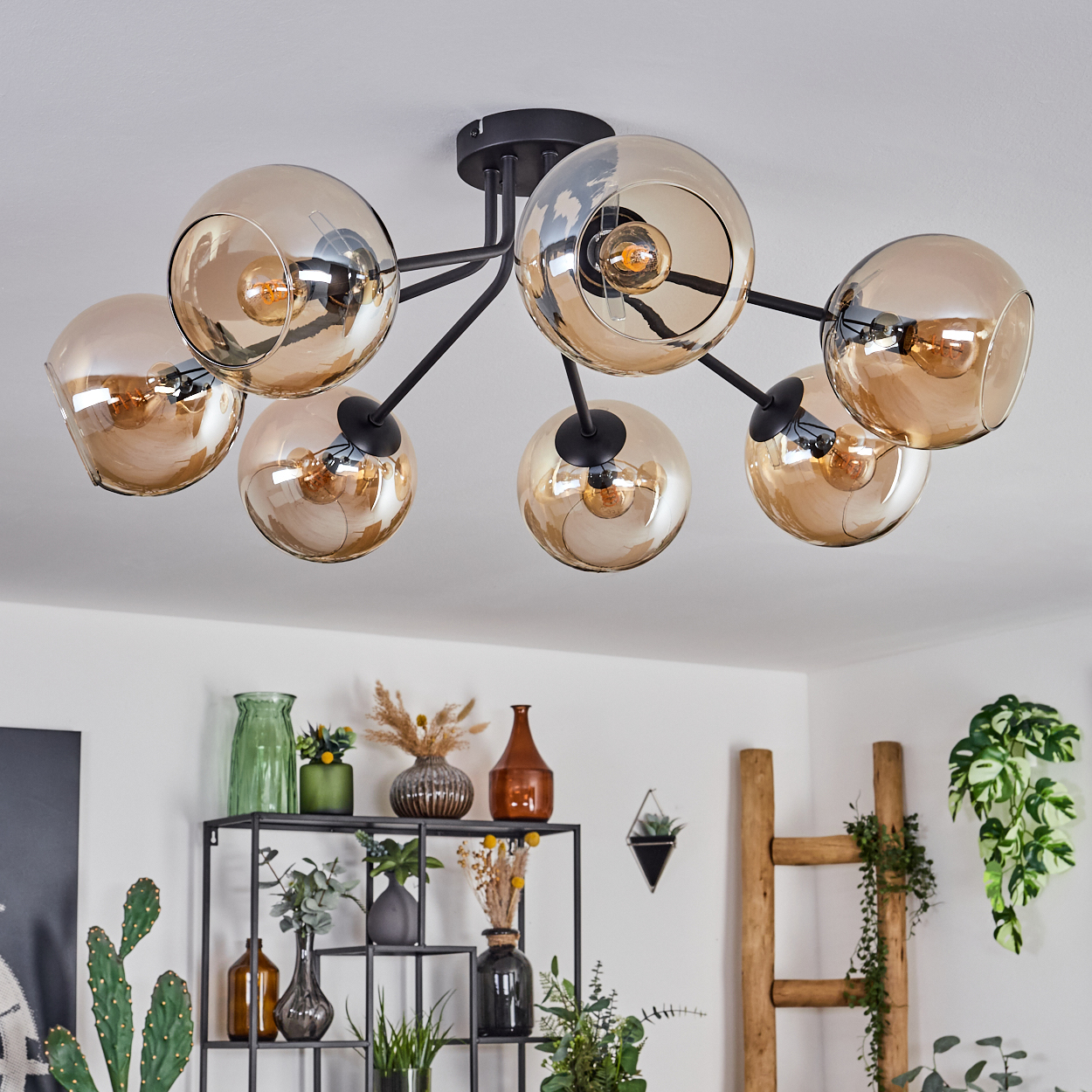 Deckenlampe mit sieben bernsteinfarbenen Glaskugeln. Schwarzer Rahmen. Pflanzen im Hintergrund.