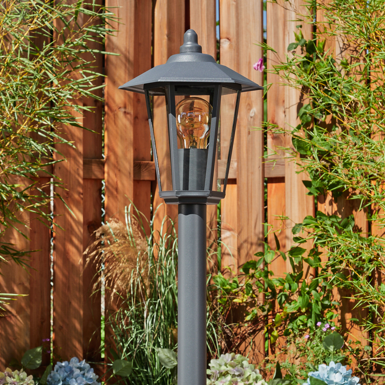 Eine graue Außenlampe mit einer leuchtenden Glühbirne steht in einem Garten vor einem Holzzaun und Grün.