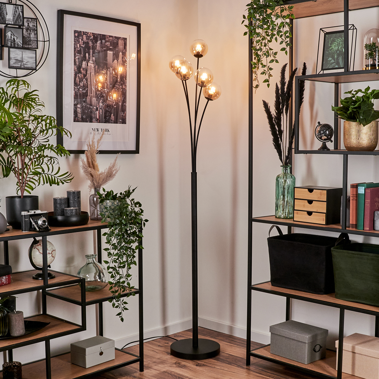 Eine schwarze Stehlampe mit fünf bernsteinfarbenen Glaskugeln neben einem Bücherregal mit Dekoration und Pflanzen.