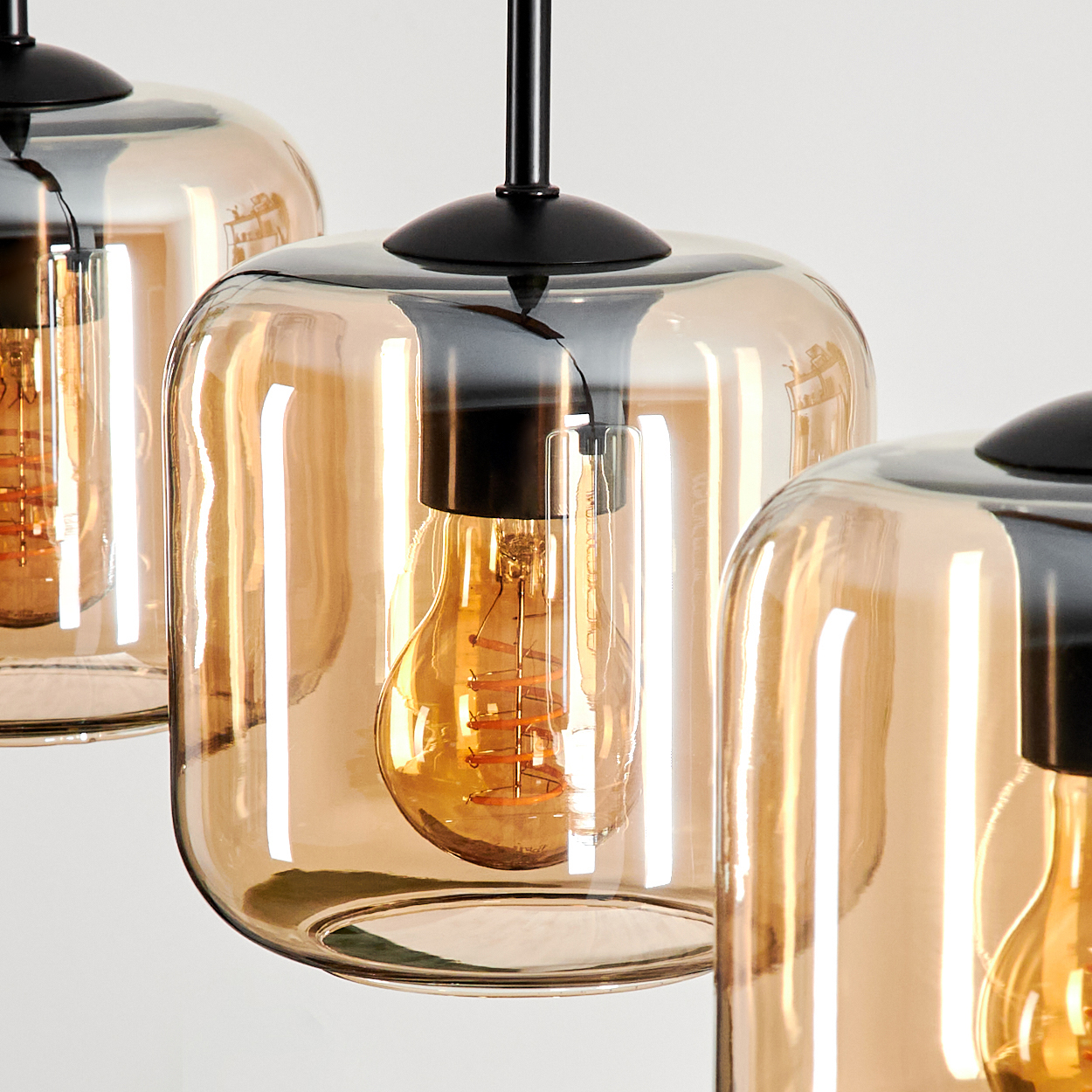 Nahaufnahme von drei bernsteinfarbenen Glas-Pendelleuchten mit Edison-Glühbirnen, schwarzen Armaturen vor Weiß.