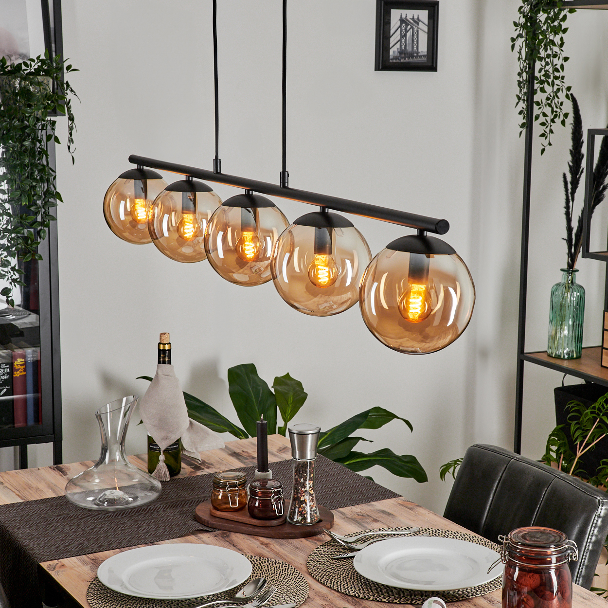 Ein Esstisch mit einer schwarzen linearen Lampe mit bernsteinfarbenen Glasschirmen, gedeckt mit Tellern, Gläsern und Kerzen.