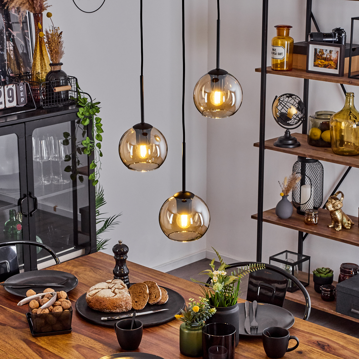 Esstisch mit Essen und drei Pendelleuchten mit bernsteinfarbenen Glasschirmen. Regale im Hintergrund.