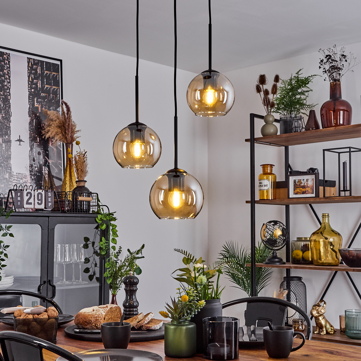 Esstisch mit Essen und drei Pendelleuchten mit bernsteinfarbenen Glasschirmen. Regale im Hintergrund.