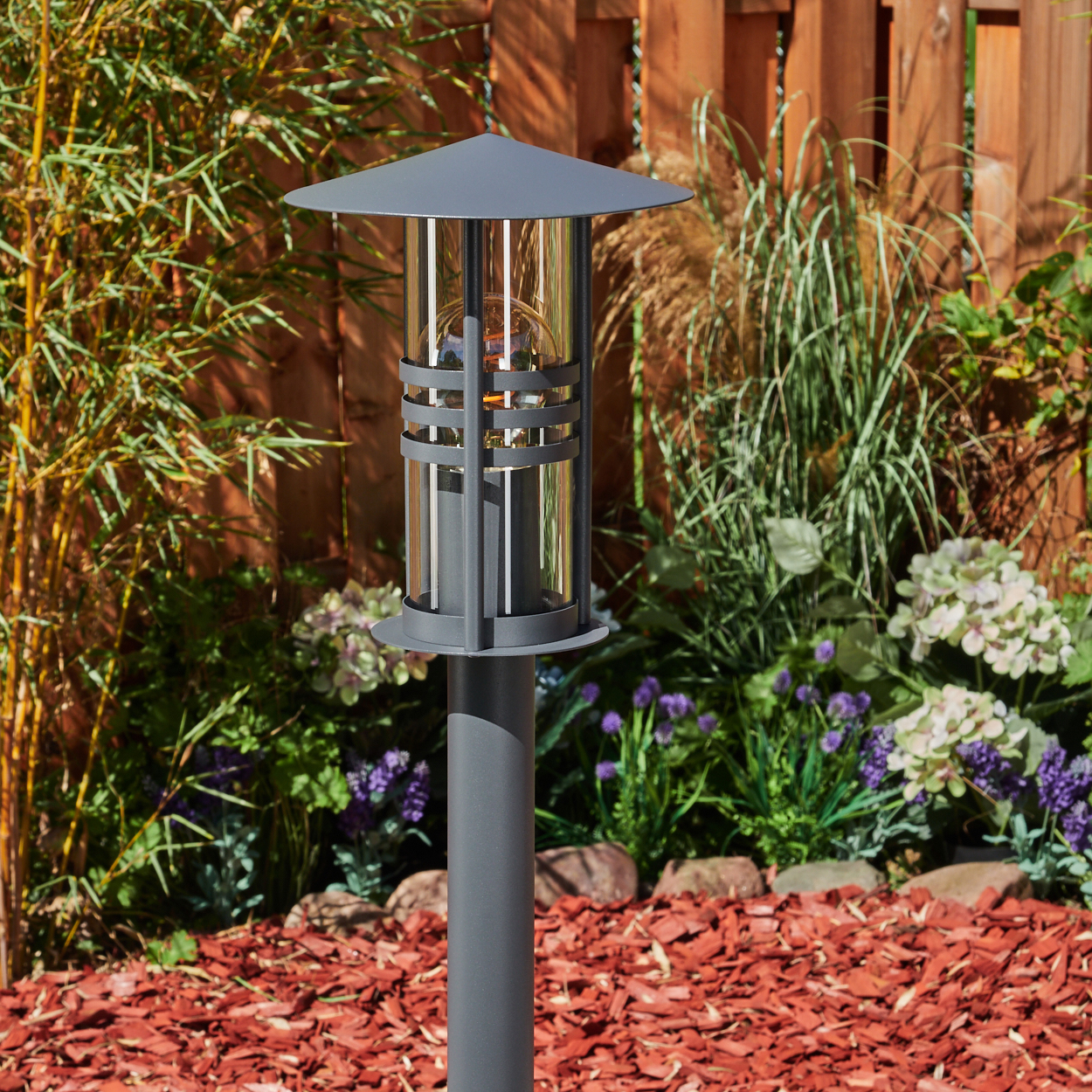 Eine graue Außenlampe mit zylindrischem Körper und klarer Glaseinfassung, in einem Garten mit Pflanzen aufgestellt.