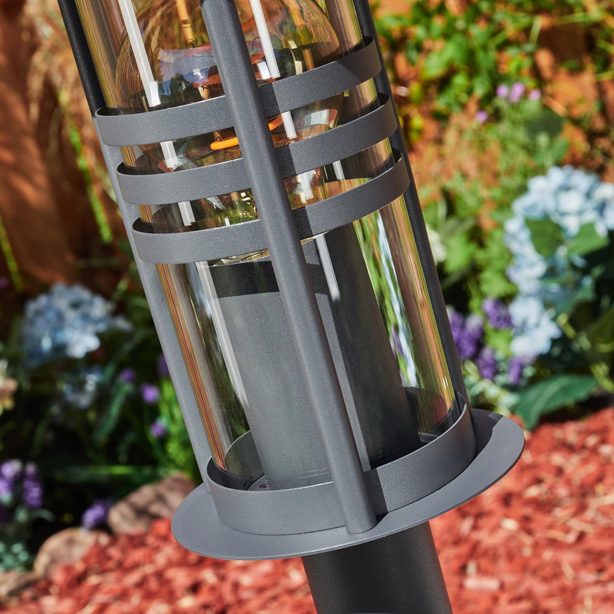 Nahaufnahme einer grauen Außenlampe mit klarer Glaseinfassung und freiliegender Glühbirne, in einer Gartenszene.