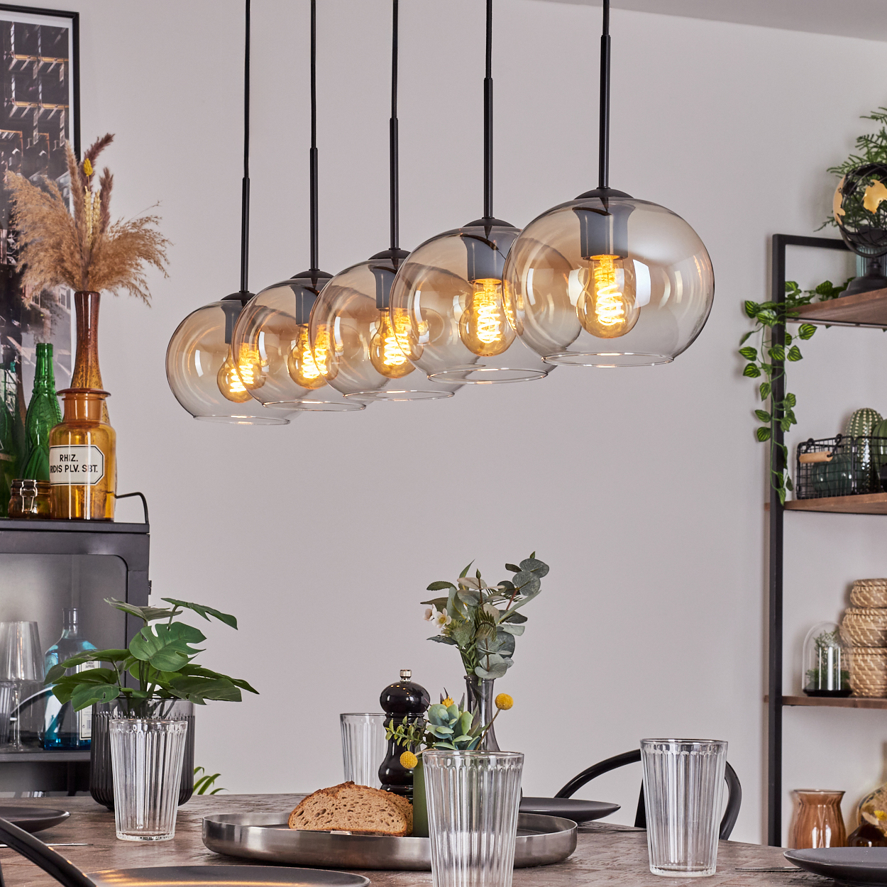 Esszimmer mit sechs Pendelleuchten, Tisch mit Gläsern und Brot gedeckt und Dekorationsartikeln auf Regalen.