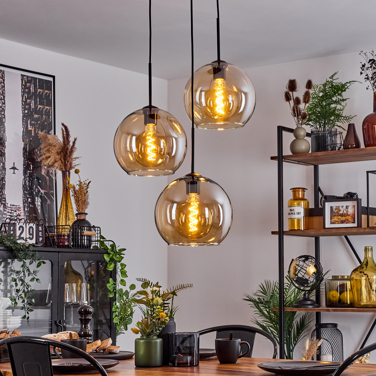Esszimmer mit einem langen Tisch und einer Deckenleuchte mit drei bernsteinfarbenen Glaskugelschirmen, mit schwarzem Metallrahmen.