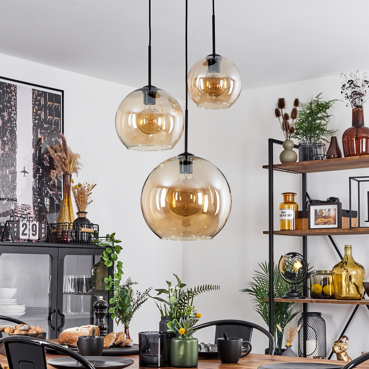 Esszimmer mit drei bernsteinfarbenen Glaskugelleuchten. Tisch mit Dekoration. Schwarzer Metallrahmen und Regale.