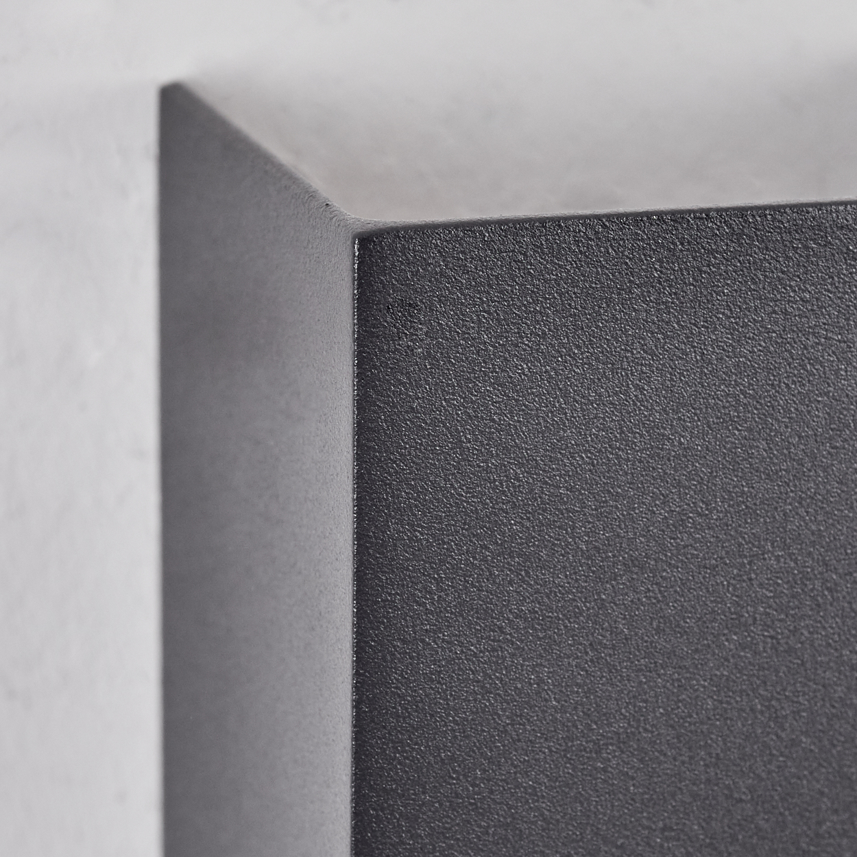 Nahaufnahme einer dunkelgrauen, strukturierten Ecke einer Box vor weißer Wand, Oberflächendetails.
