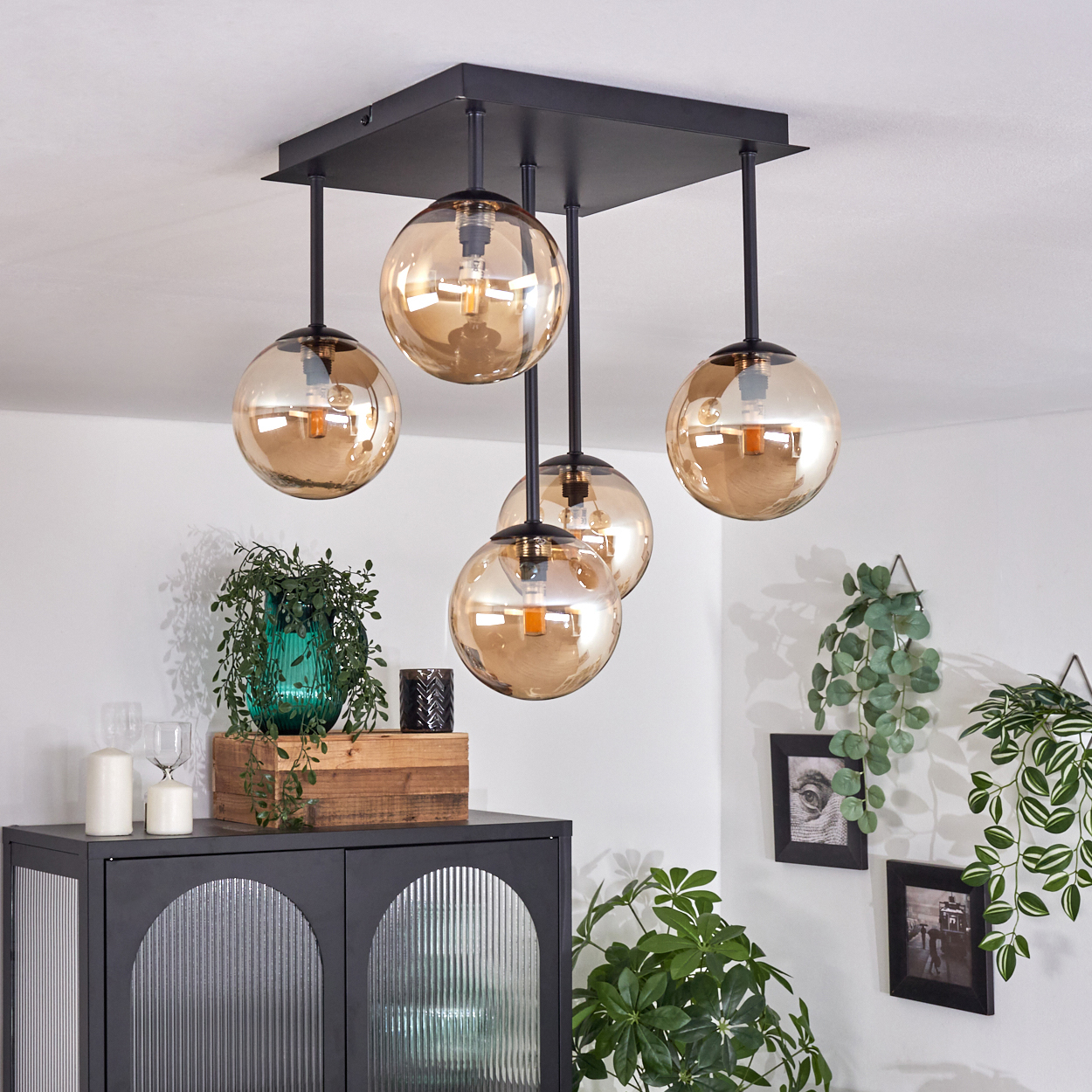 Deckenlampe mit fünf bernsteinfarbenen Glaskugeln. Schwarzer Rahmen und Aufhängestangen. Raum mit Dekoration im Hintergrund.