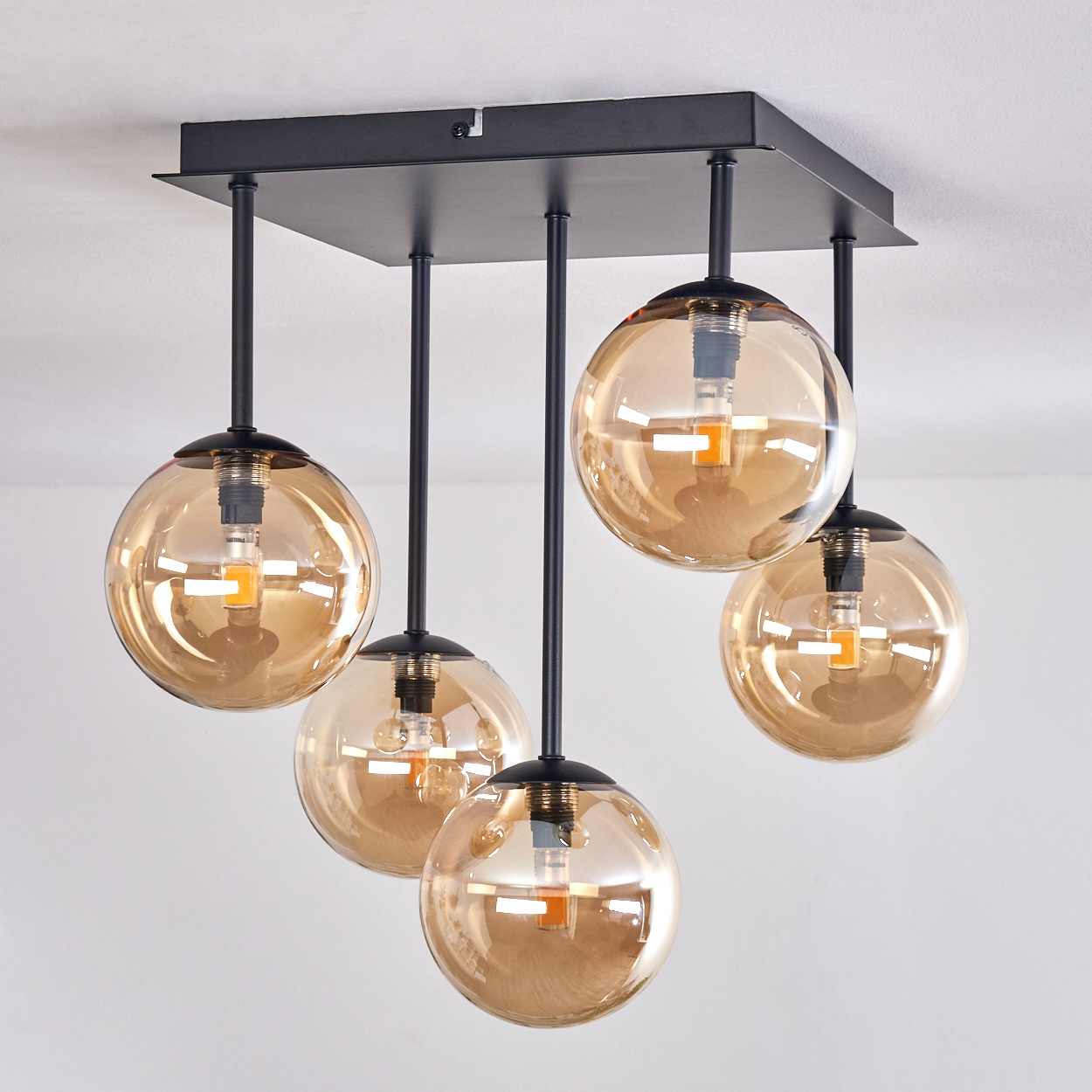 Deckenlampe mit fünf bernsteinfarbenen Glaskugeln. Schwarzer Rahmen und Aufhängestangen. Weißer Hintergrund.