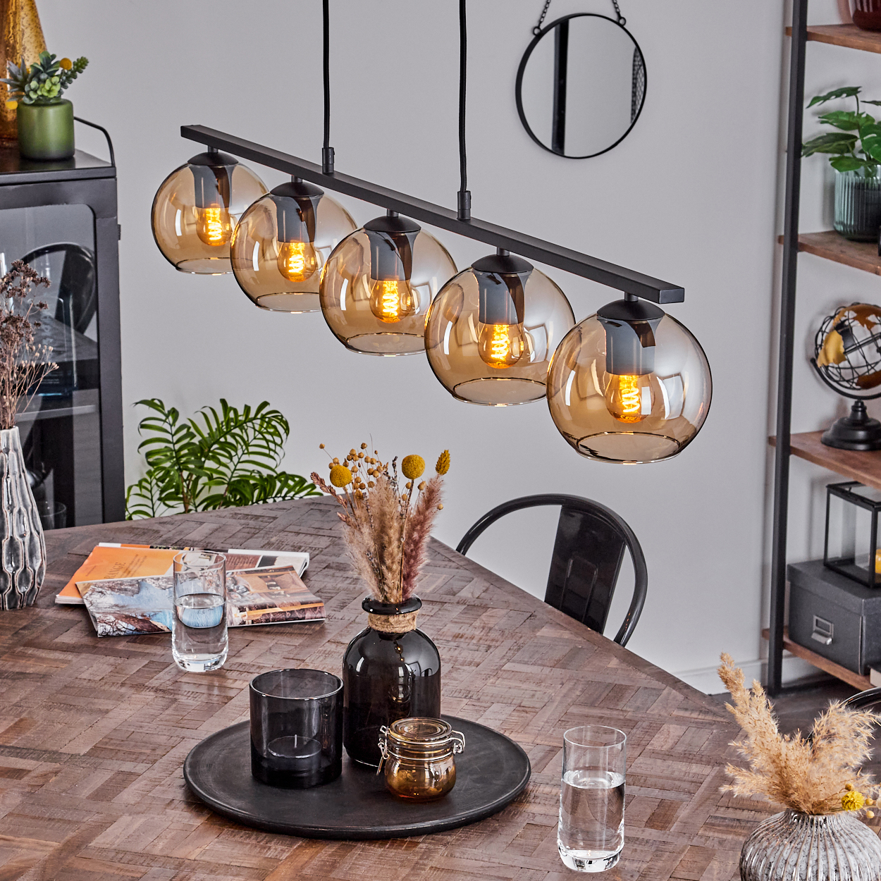 Eine schwarze lineare Pendelleuchte mit fünf bernsteinfarbenen Glaskugeln hängt über einem Holztisch mit Tablett und Deko.