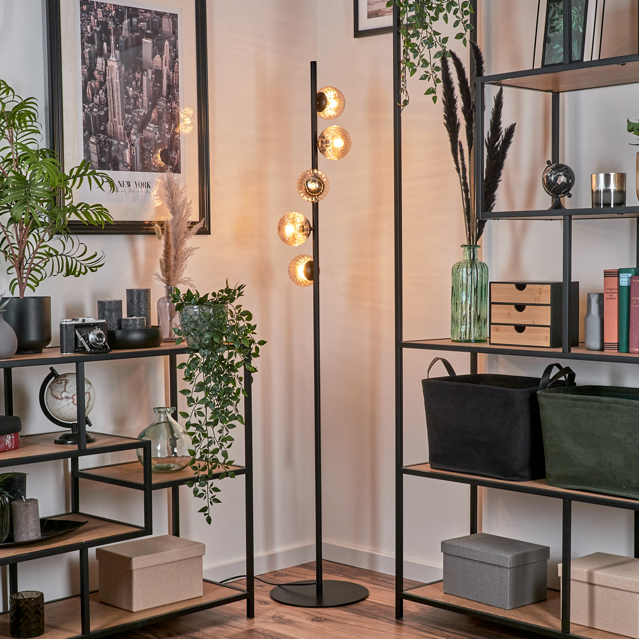 Eine schwarze Stehlampe mit fünf bernsteinfarbenen Glaskugeln steht in einer Ecke, mit Pflanzen und Regalen drumherum.