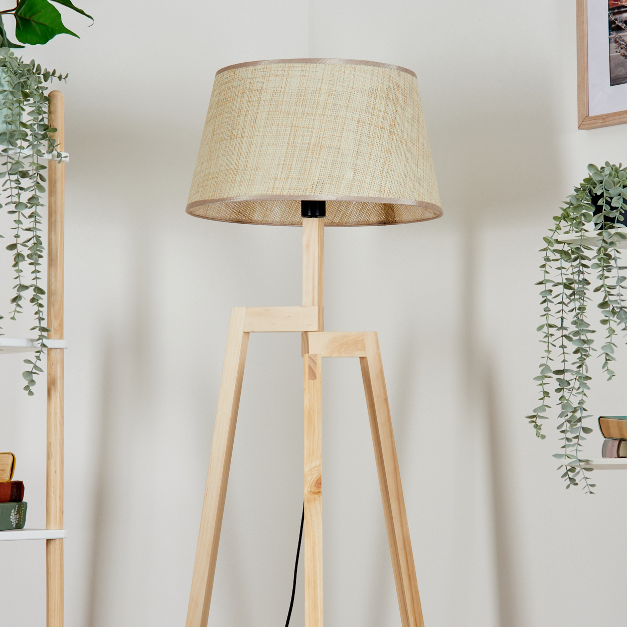 Stehlampe mit Holzbeinen, beigem Stoffschirm, Pflanzen im Hintergrund.