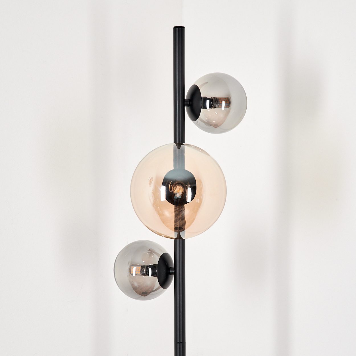Eine schwarze Lampe mit drei Kugelschirmen, einer bernsteinfarbenen und zwei silbernen, vor einer weißen Wand.