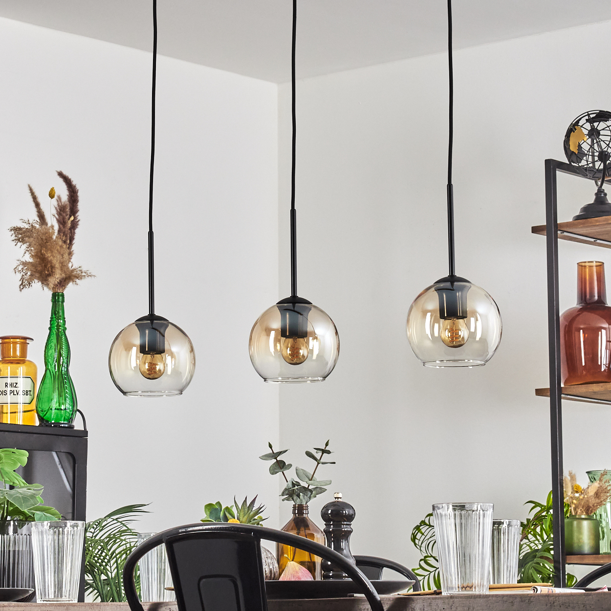 Drei bernsteinfarbene Glas-Hängeleuchten hängen über einem Tisch mit Gläsern und Deko.