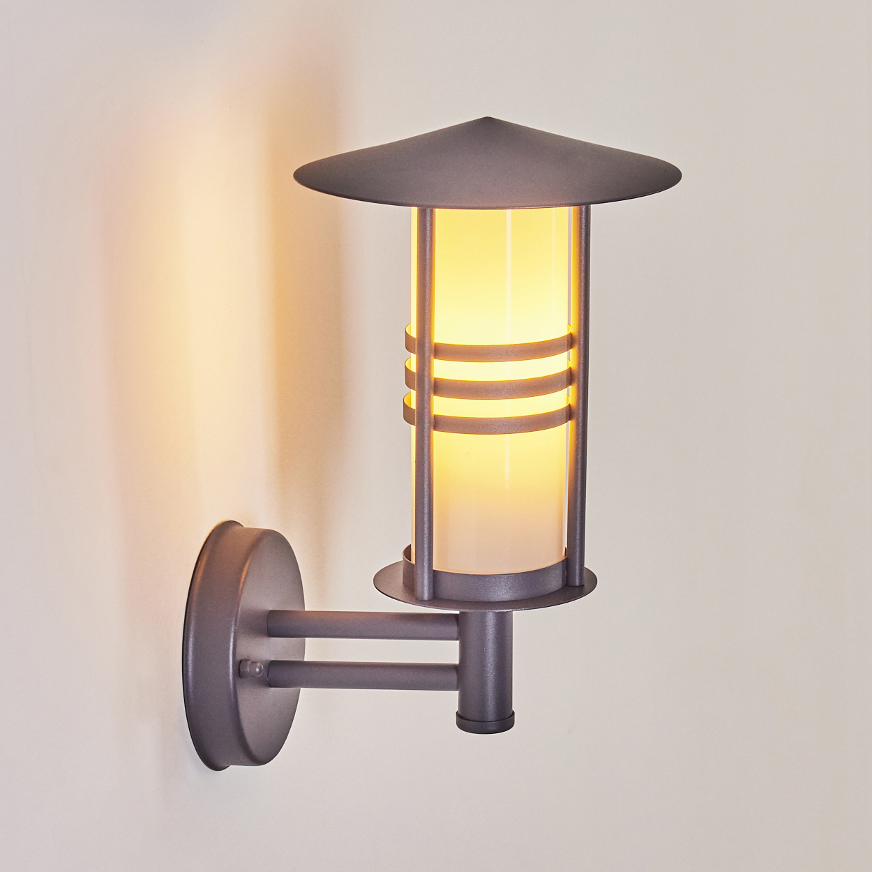 Beleuchtete Außenwandleuchte. Das Licht scheint durch den zylindrischen Diffusor. Hintergrund ist eine weiße Wand.
