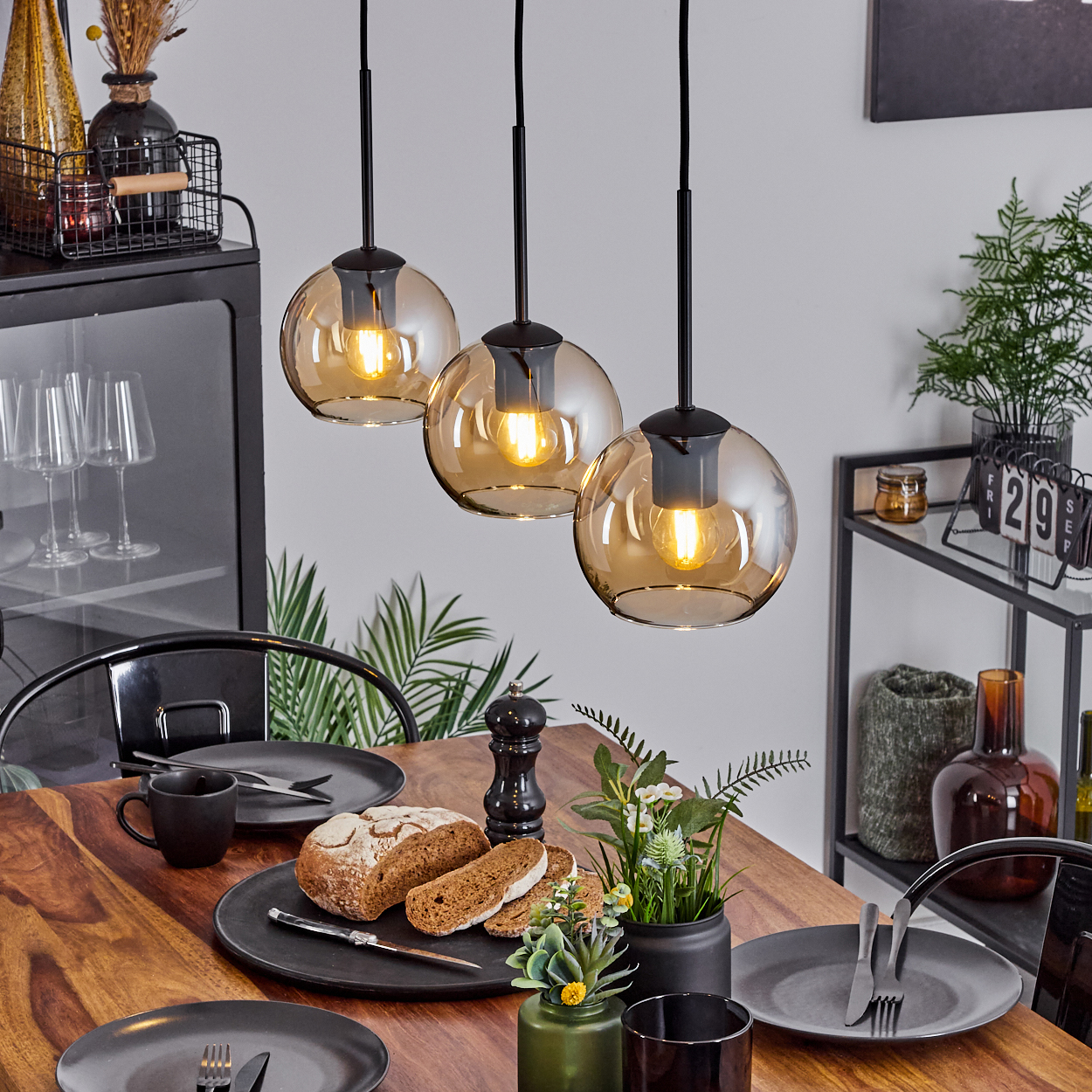 Esszimmer mit drei Pendelleuchten mit bernsteinfarbenen Glaskugeln. Holztisch, Brot und Deko sind vorhanden.