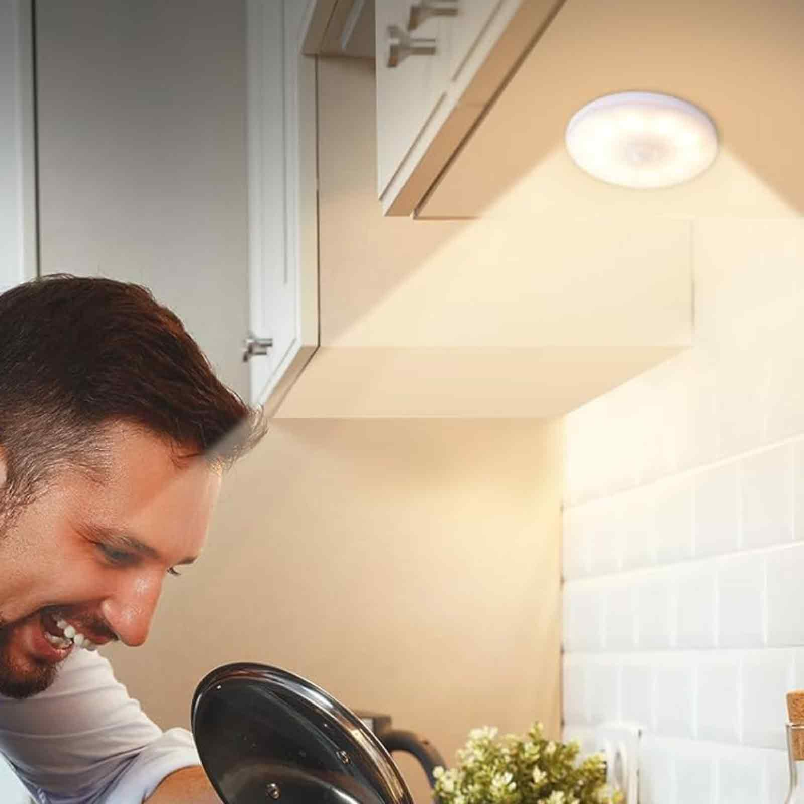 Ein Mann lächelt eine Leuchte in einer Küche an. Es ist eine kleine, runde, weiße LED-Leuchte.