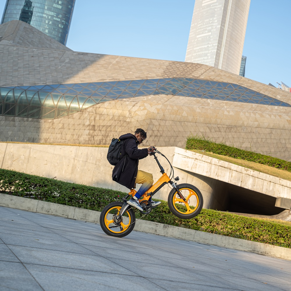 Mann in schwarzer Jacke fährt auf einem orangefarbenen Fahrrad, Vorderrad angehoben, vor einem modernen Gebäude.