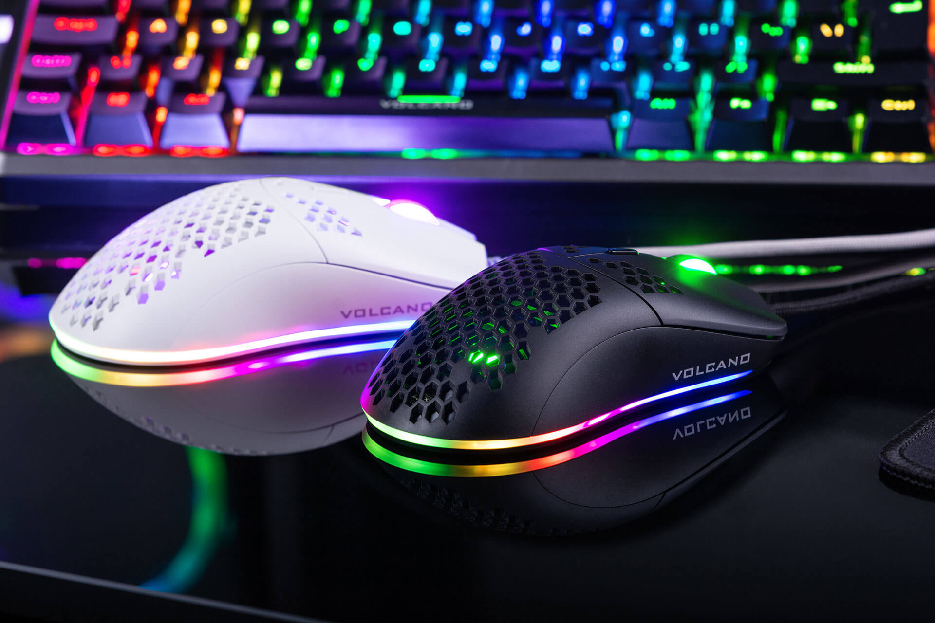 Dwie myszy do gier, biała i czarna, z oświetleniem RGB, leżą na czarnej powierzchni przed klawiaturą.