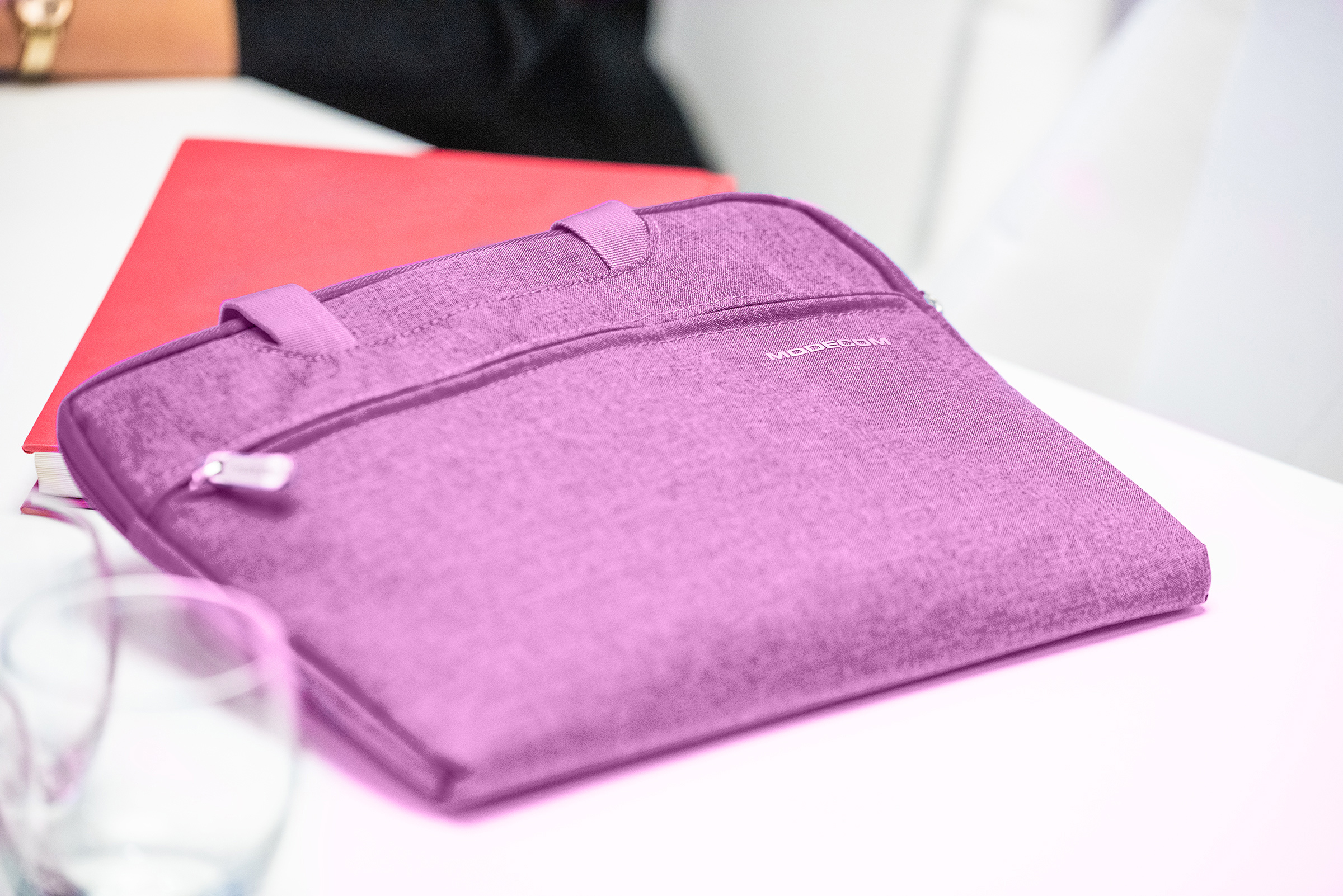 Fioletowa torba na laptopa na stole, z czerwoną książką za nią. Torba ma zamek błyskawiczny i napis 'MODECOM'.