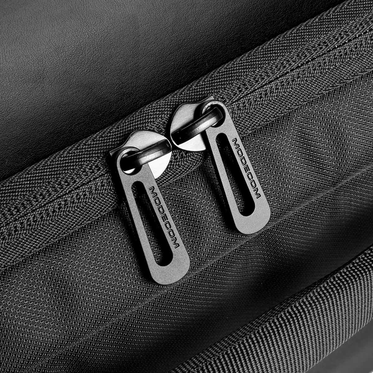 Zbliżenie czarnych uchwytów zamków błyskawicznych z napisem "MODECOM" na czarnym tle materiału.