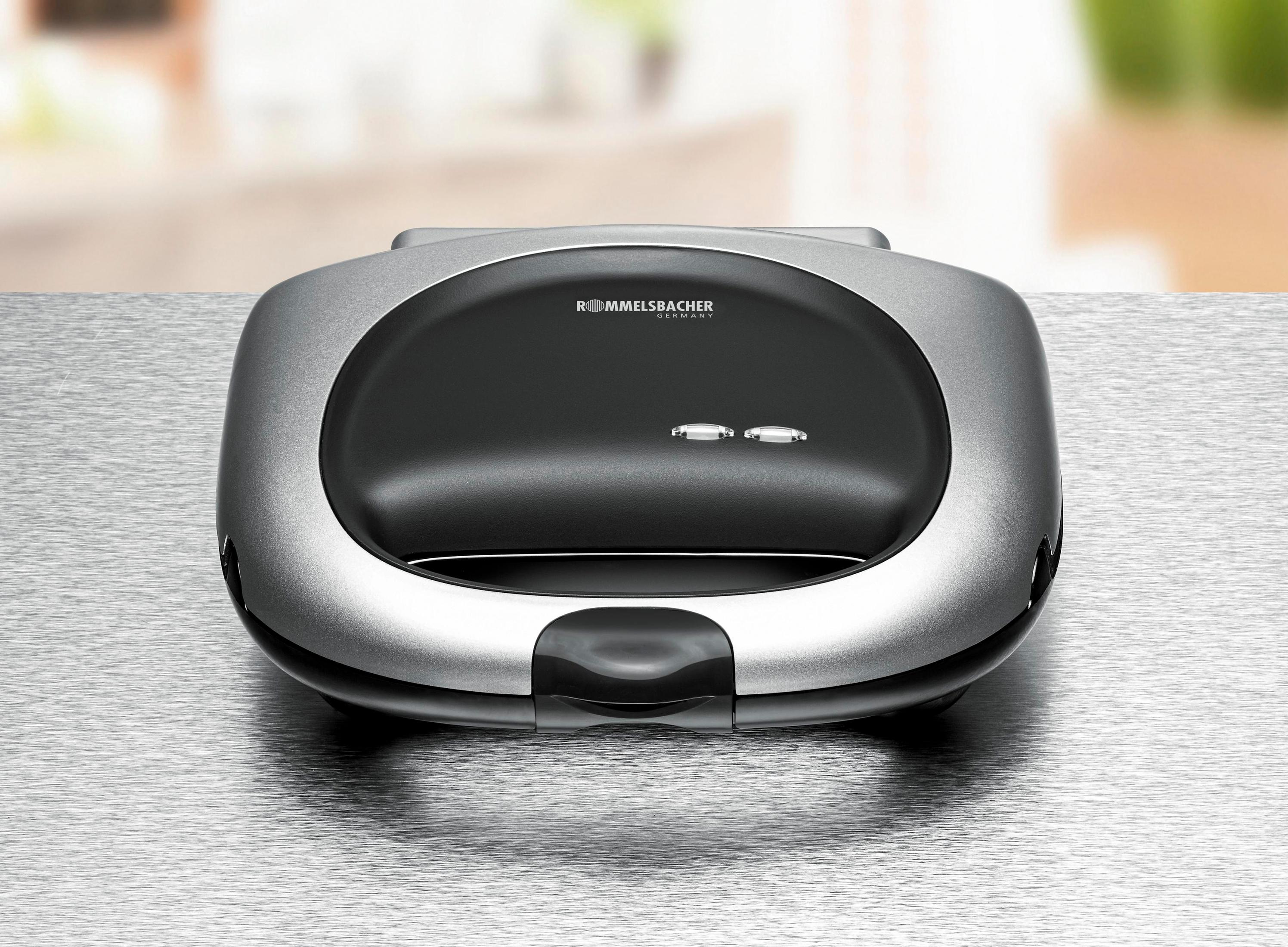 Srebrny opiekacz do kanapek. Czarna góra z dwoma światłami i nazwą marki. Błyszcząca powierzchnia. Na srebrnym stole.