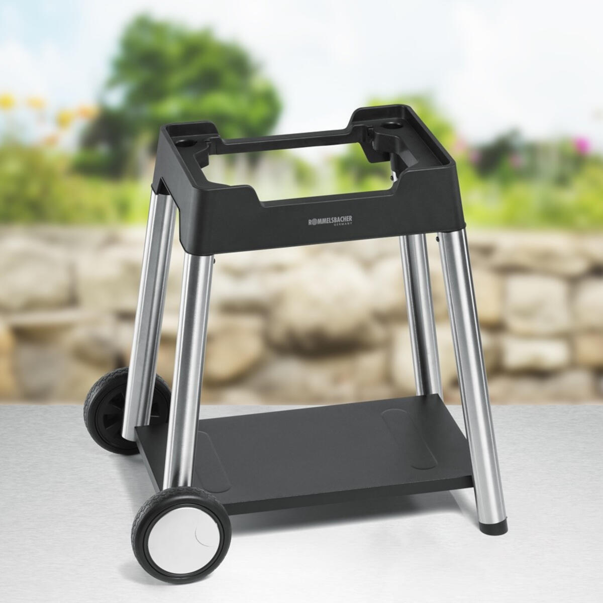 Czarny metalowy stojak na grilla z kołami. Srebrne nogi, czarny blat z marką i półka poniżej.