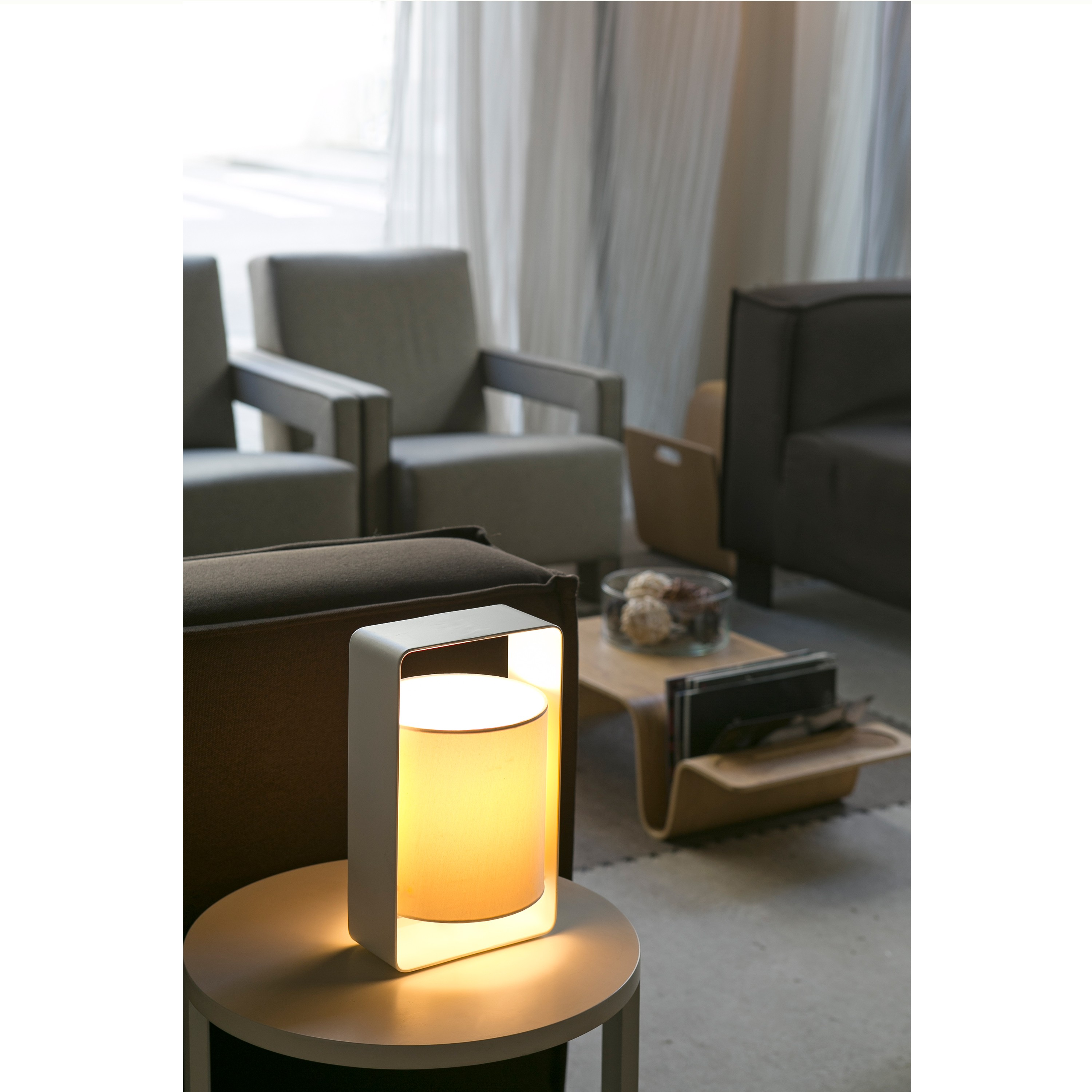 Lampada da tavolo bianca con cornice rettangolare e paralume cilindrico illuminato, su tavolo rotondo.
