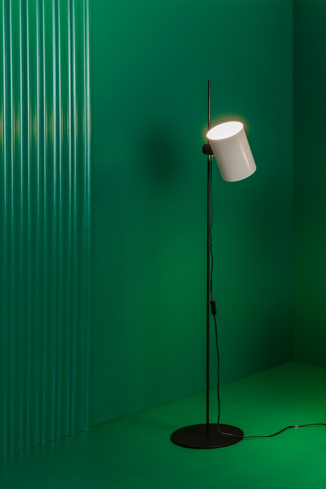 Una lampada da terra nera con paralume bianco contro un muro verde, pavimento nero.