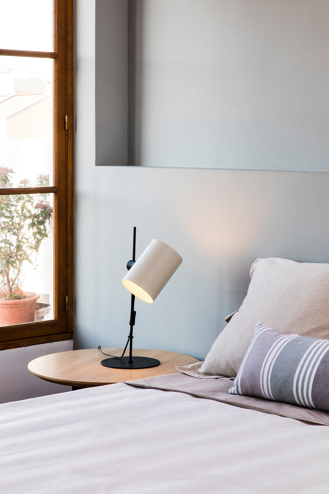 Lampada bianca su un tavolo vicino a un letto. Lo sfondo è un muro azzurro e una finestra.