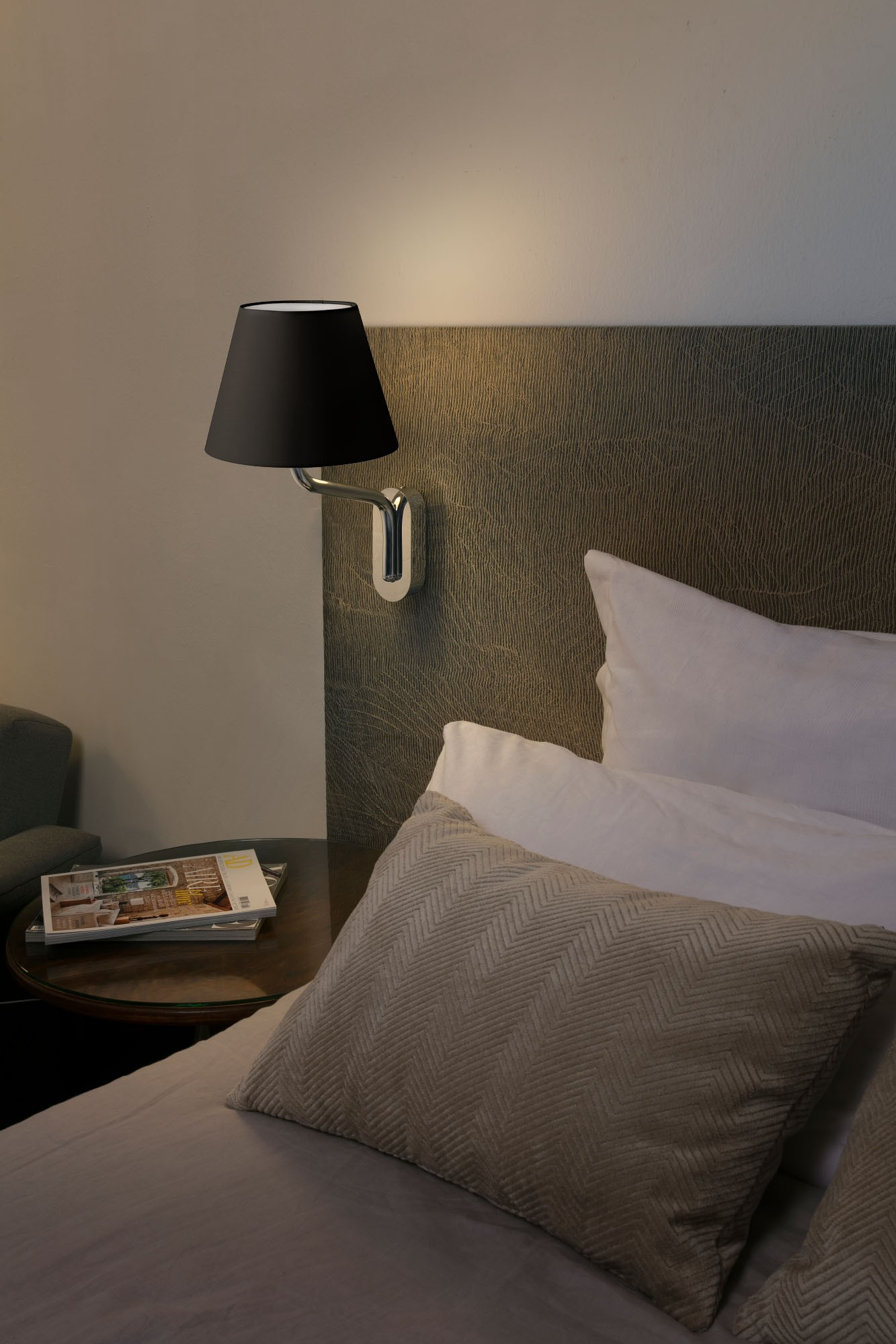 Un letto con cuscini e una lampada. Una lampada nera è sulla parete. Una rivista è sul comodino.