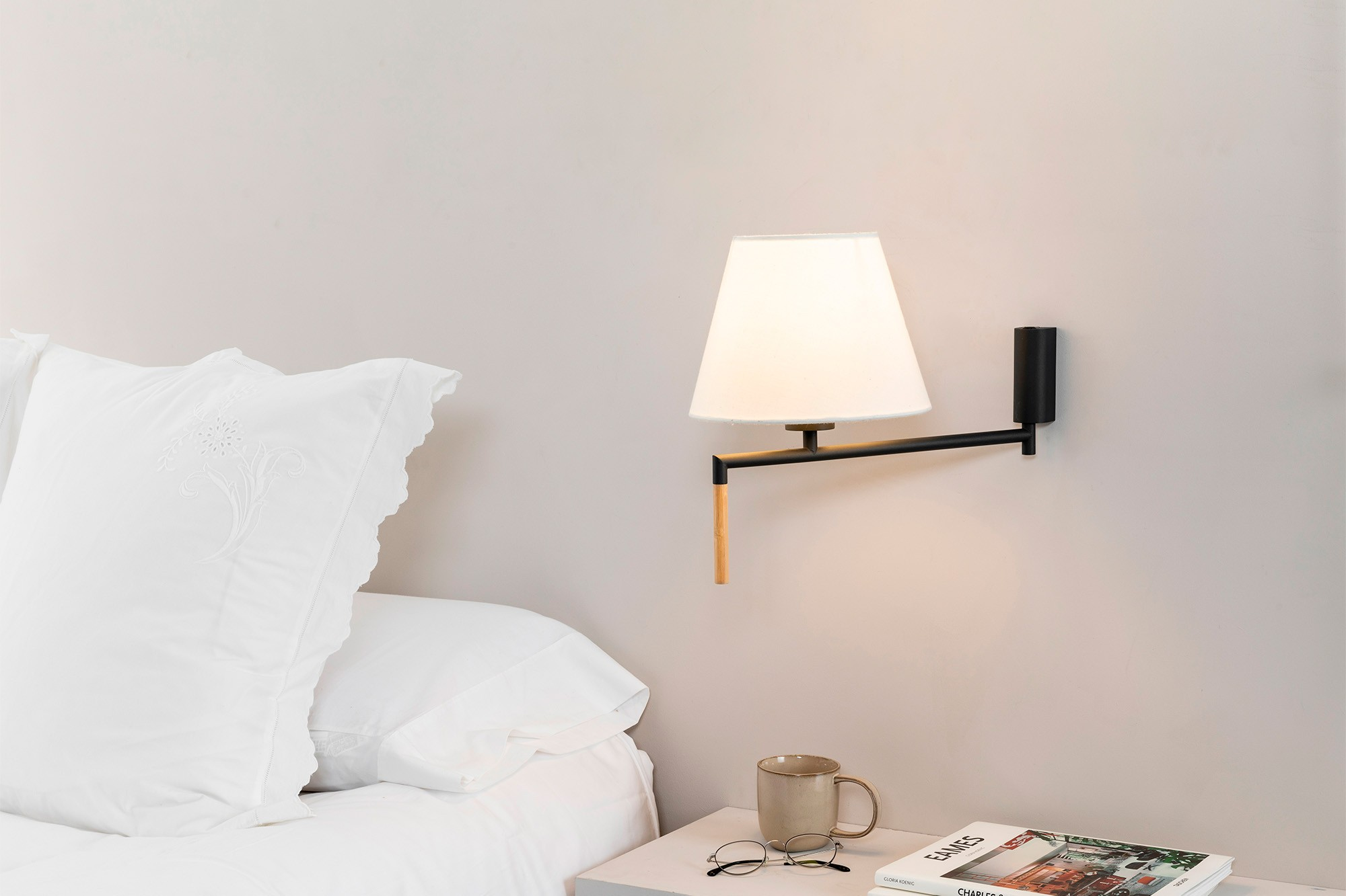 Lampada da parete con paralume bianco, accento in legno e libro su comodino con tazza.