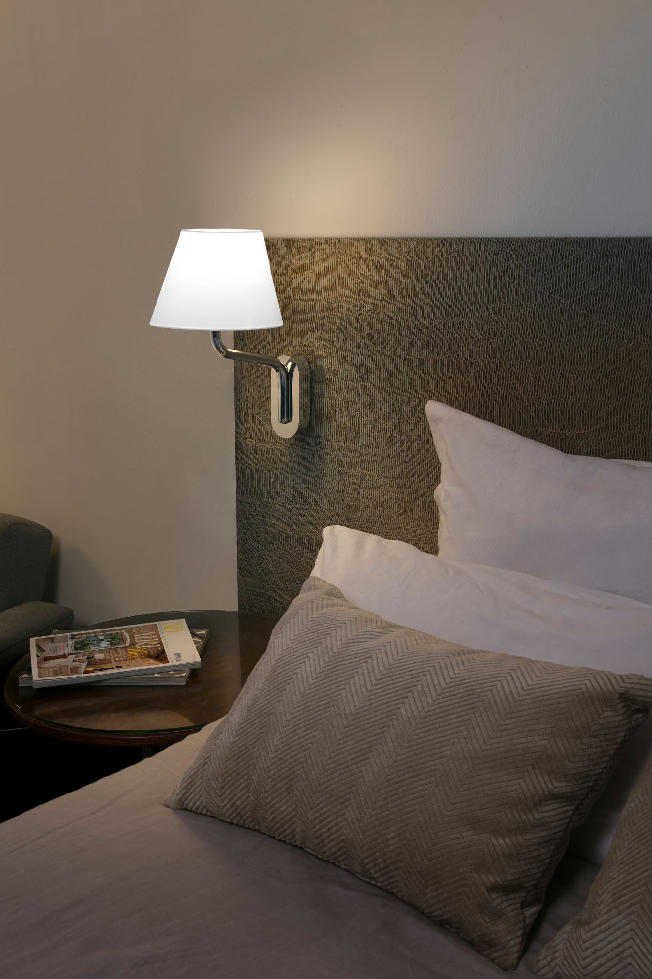 Lampada da parete con paralume bianco sopra un letto. Un libro e un tavolo in primo piano.