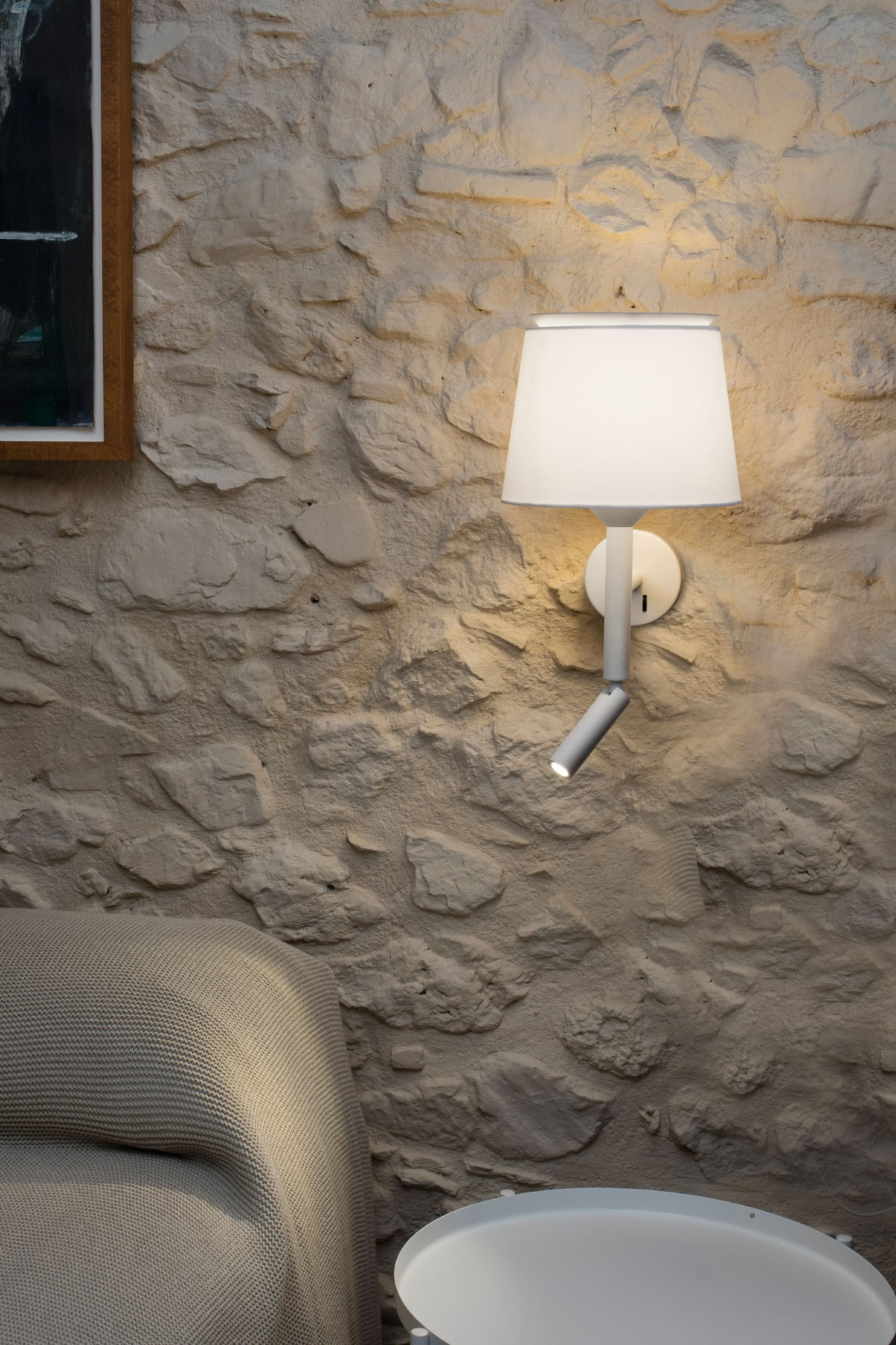 Lampada da parete con paralume bianco e piccola luce da lettura. Muro di pietra sullo sfondo.