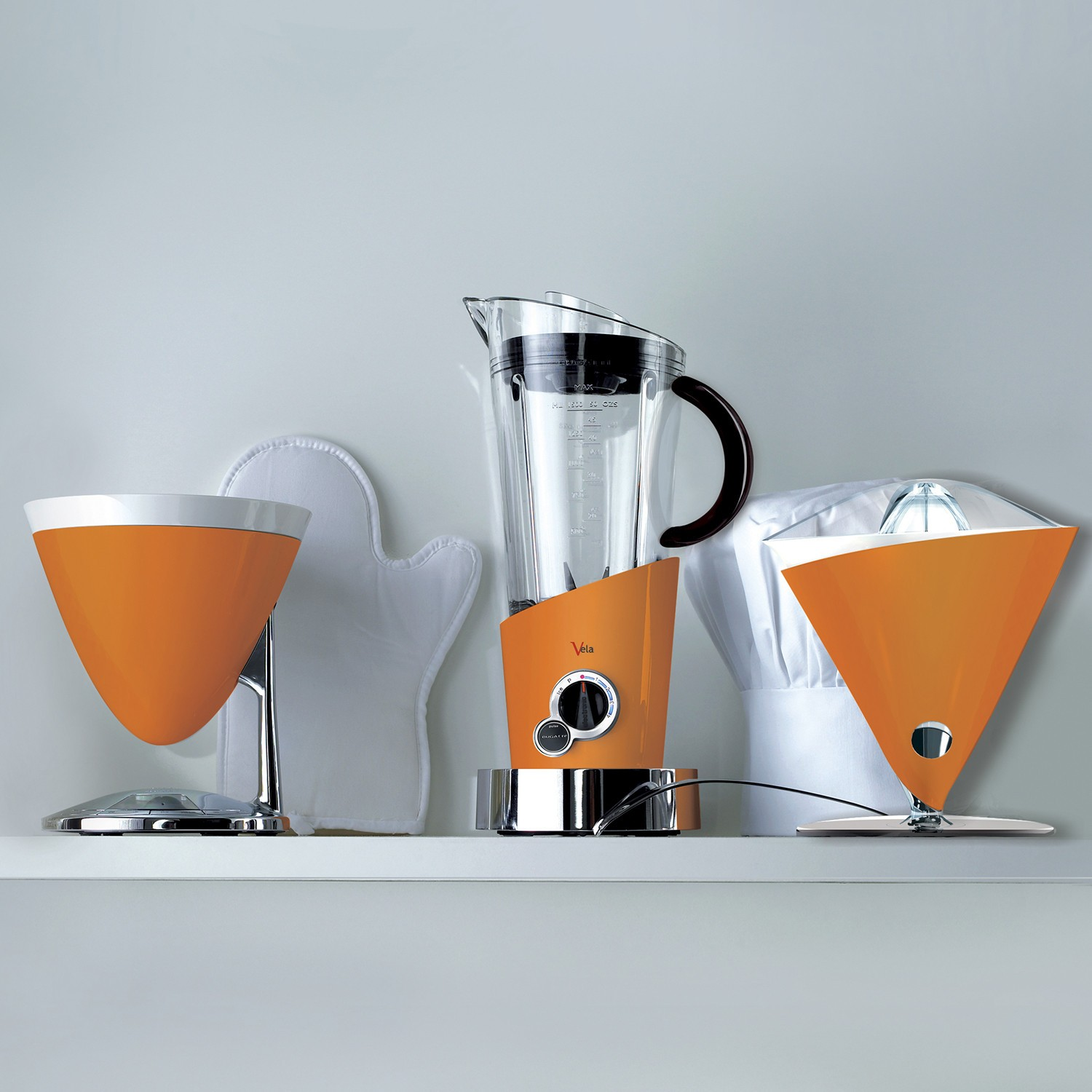 Allestimento cucina con elettrodomestici arancioni: un frullatore, uno spremiagrumi e una caffettiera. Mensola bianca.