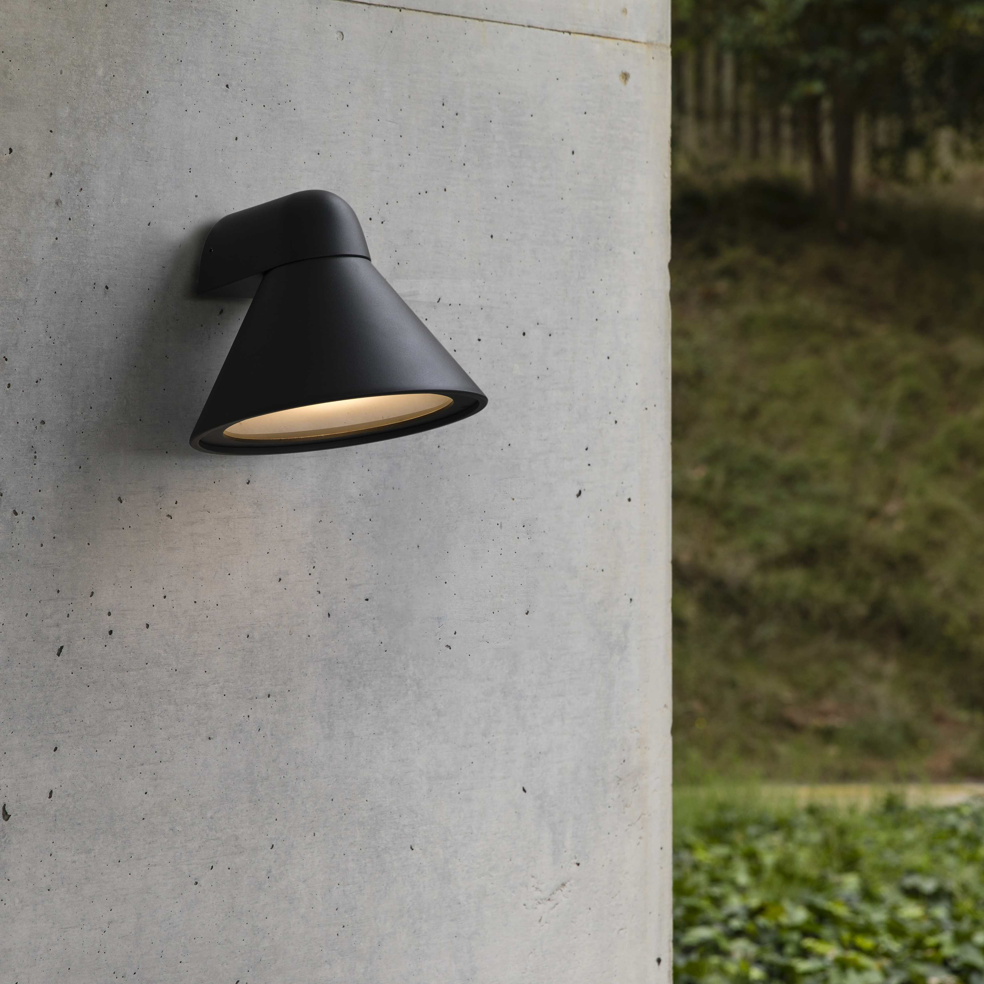 Schwarze Außenlampe an einer Betonmauer, Grün im Hintergrund.