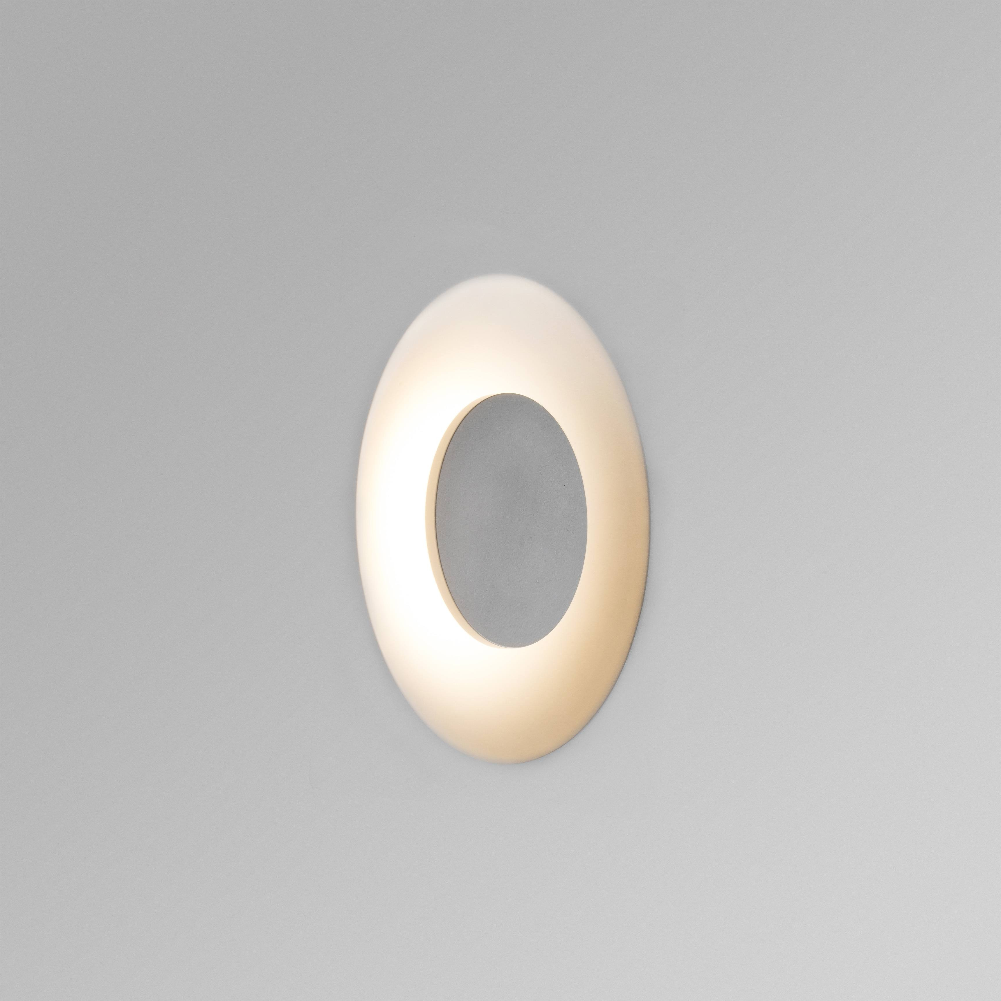 Una lampada bianca di forma ovale montata su un muro grigio. Emette luce.