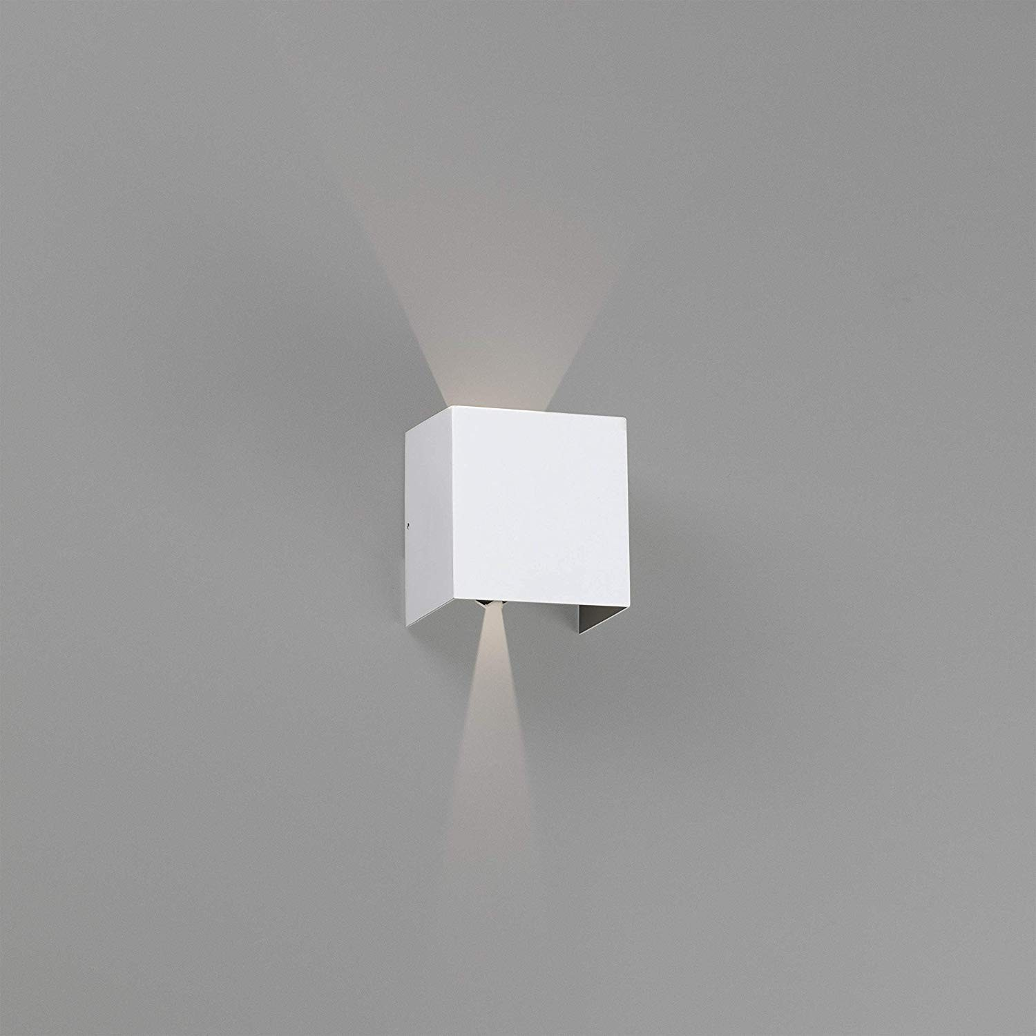 Una lampada da parete a cubo bianca su un muro grigio, che emette luce verso il basso.