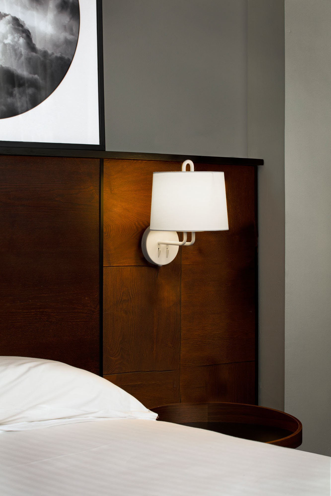 Una lampada da parete con paralume bianco vicino a un letto.