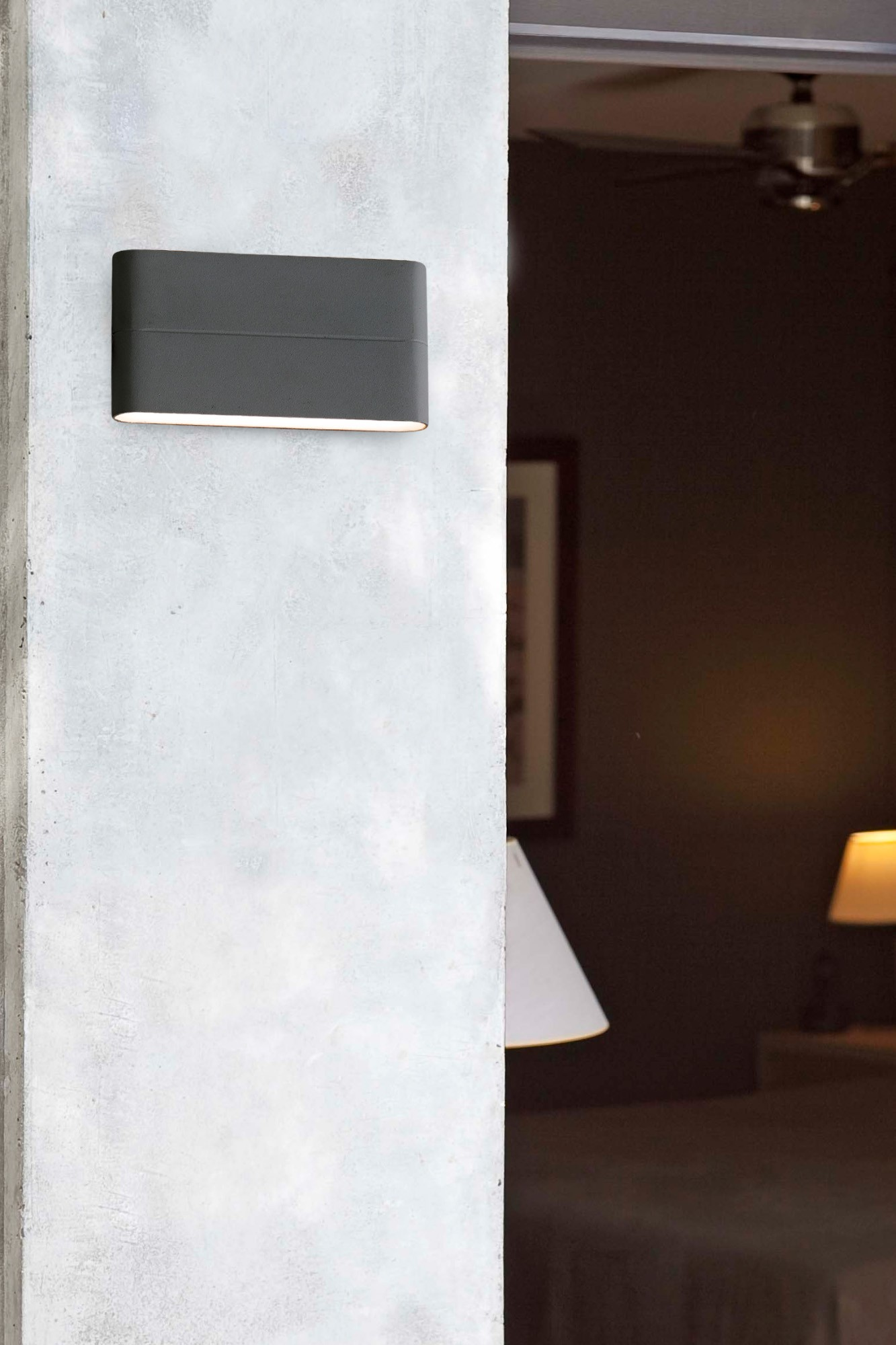 Eine graue Wand mit einer Lampe. Durch ein Glasfenster sind eine Tür und ein Raum zu sehen.