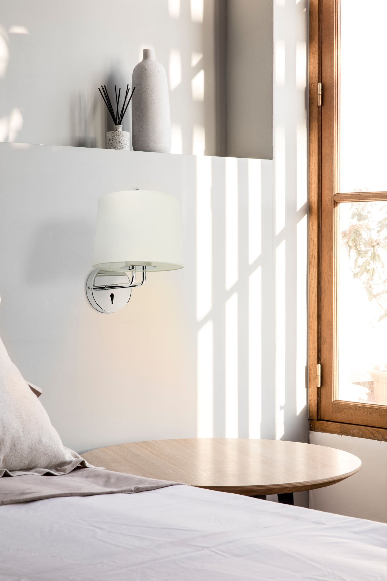 Una lampada da parete con paralume bianco vicino a una finestra.