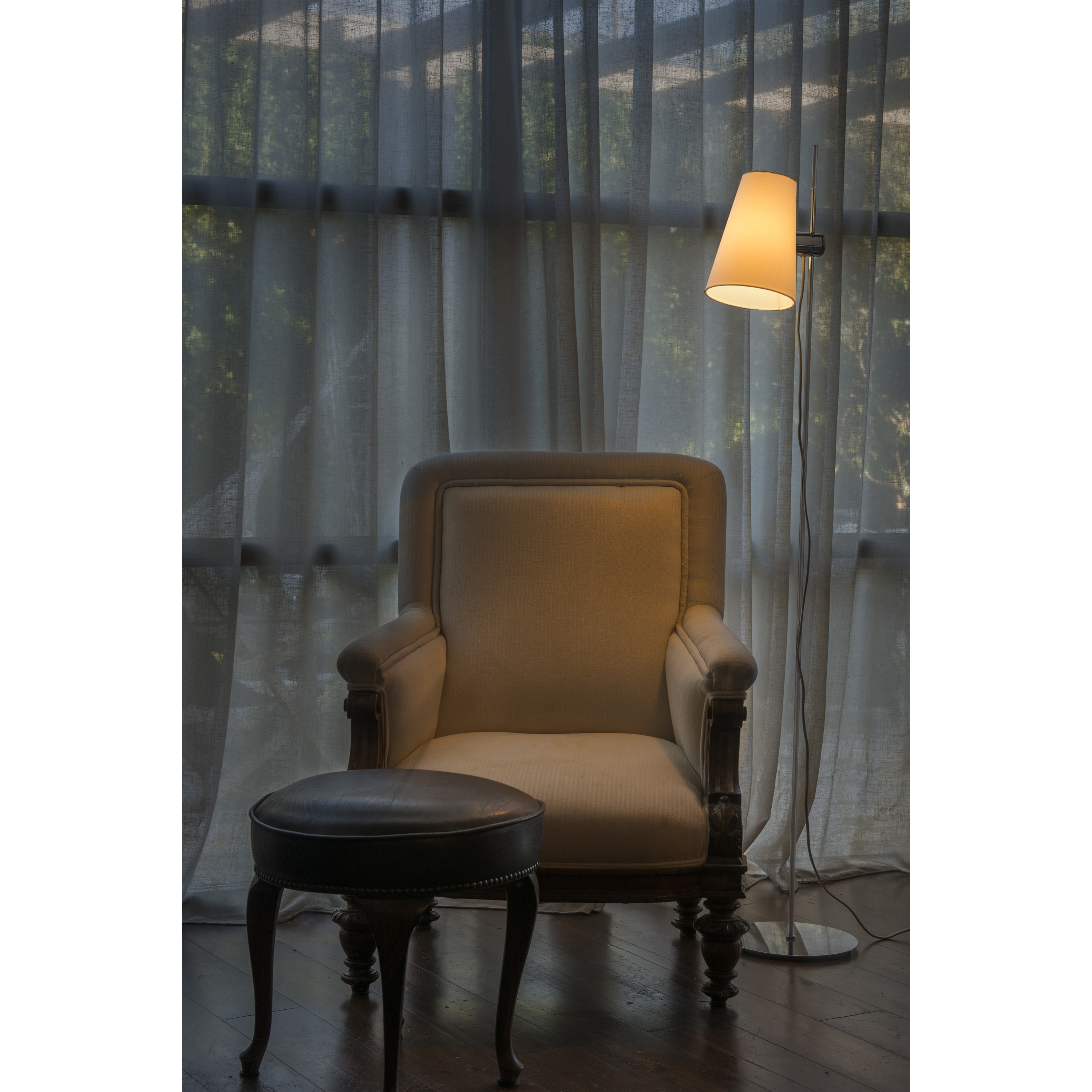 Poltrona con lampada da terra, dietro tende trasparenti. Un piccolo sgabello di fronte.