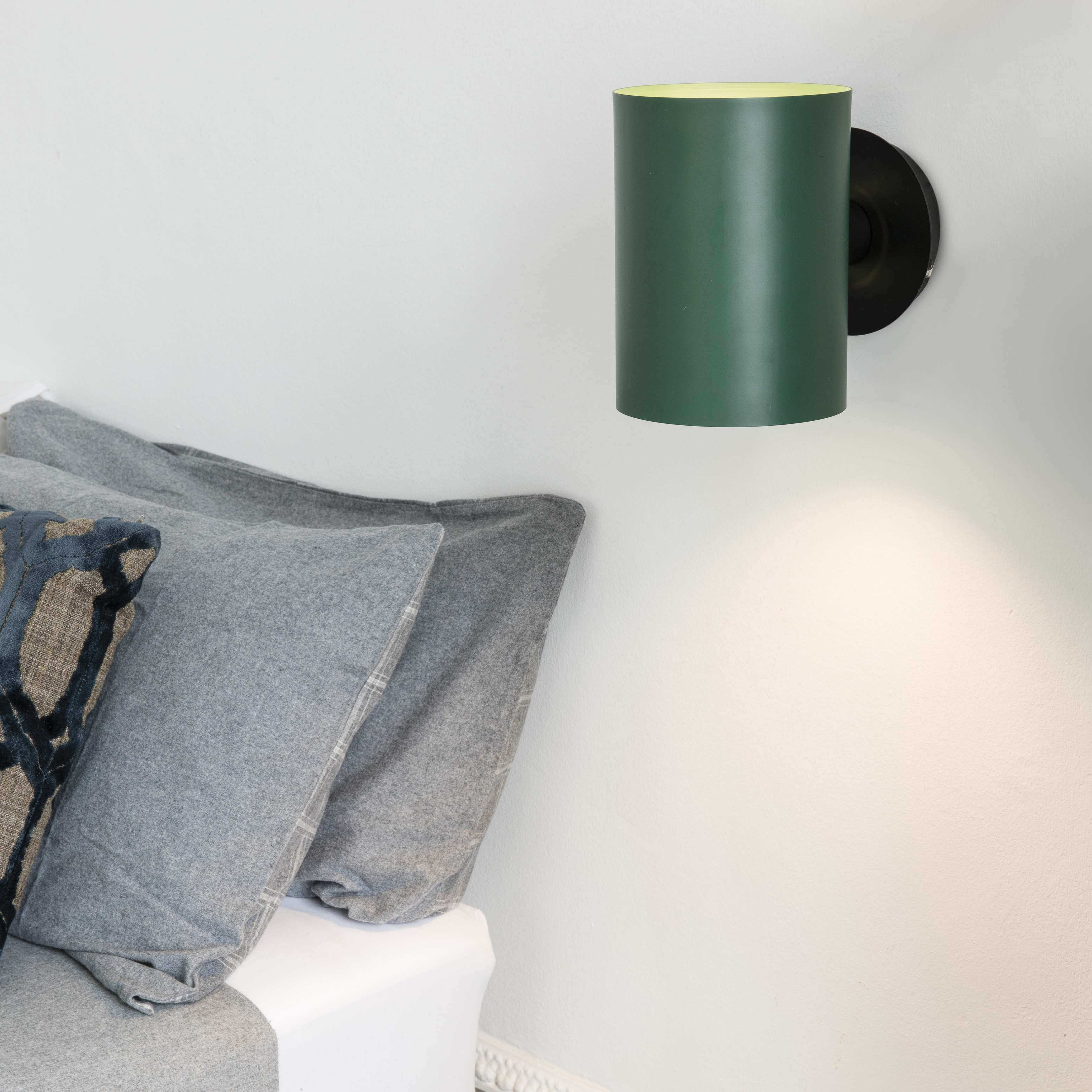 Lampada da parete cilindrica verde su parete bianca. Cuscini grigi su letto bianco.